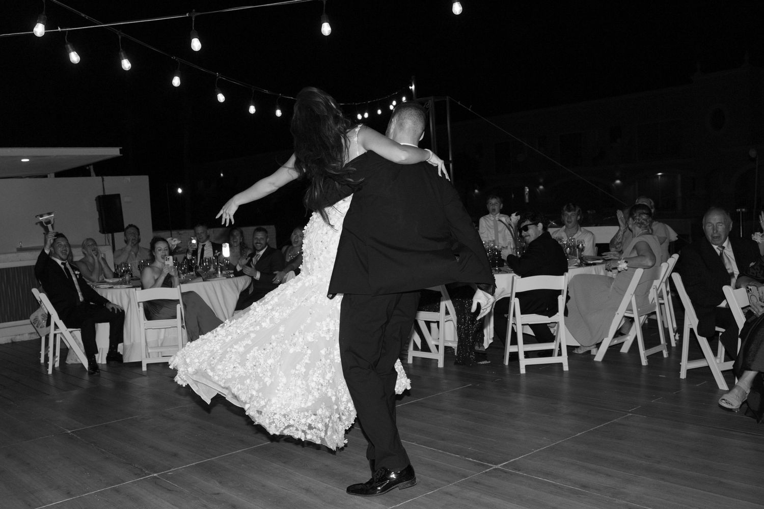 A couple shares their first dance on a wooden floor under string lights at their wedding reception.