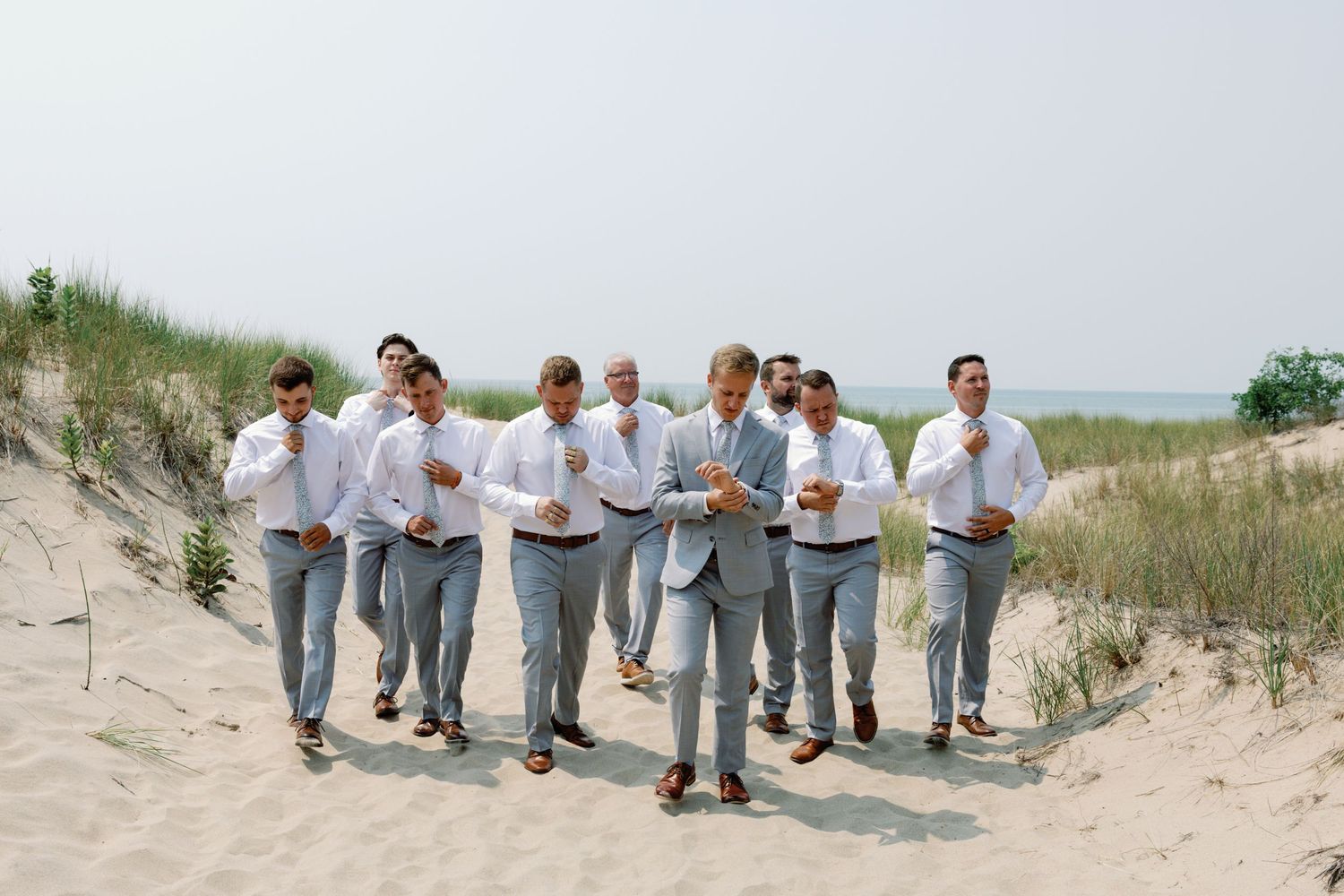 Groomsmen in light gray suits posing playfully on beach sand dunes.