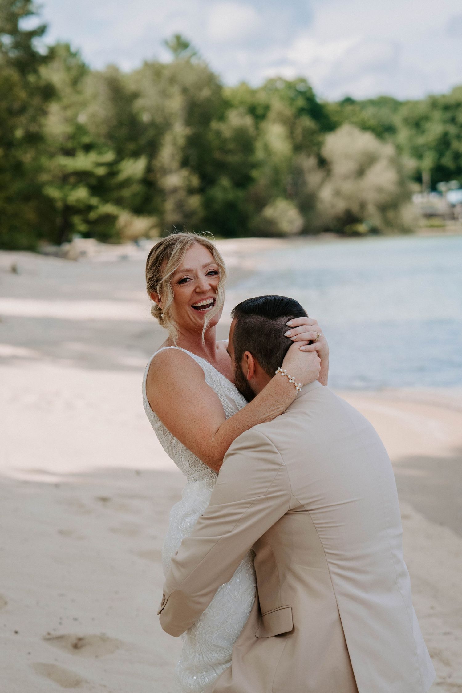 Couple in wedding attire embrace on a sunny beach with water and trees in background.