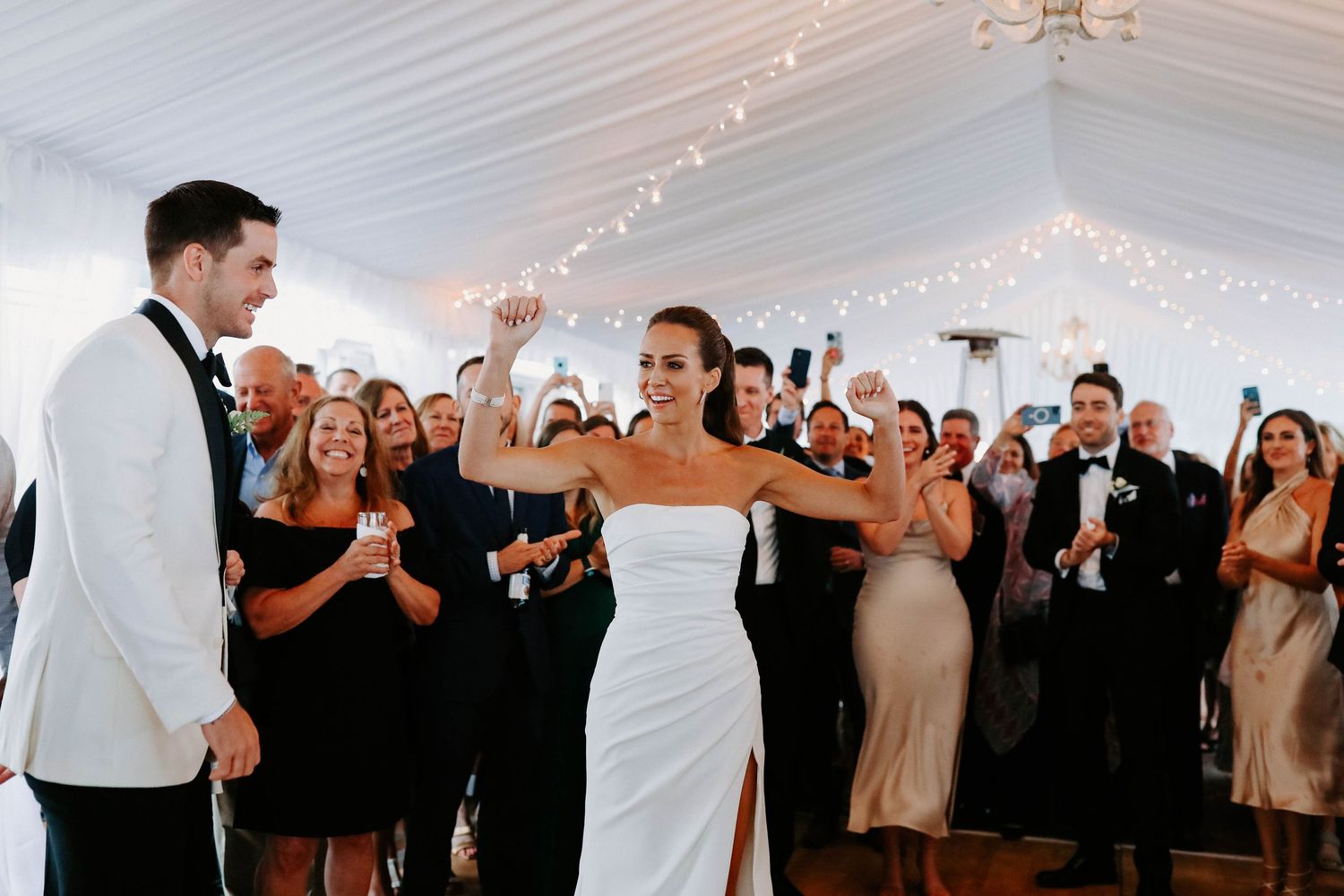 Wedding party dancing and celebrating under string lights in an elegant white tent reception space.