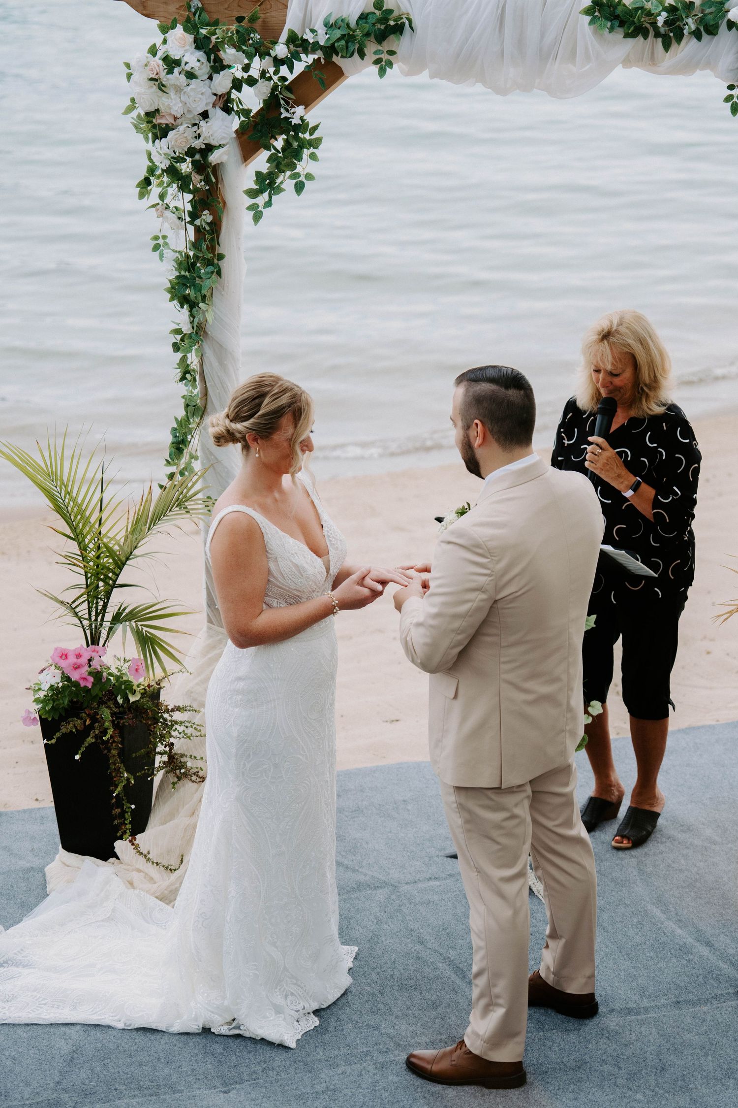 A wedding ceremony under a floral arch by the water as rings are exchanged.