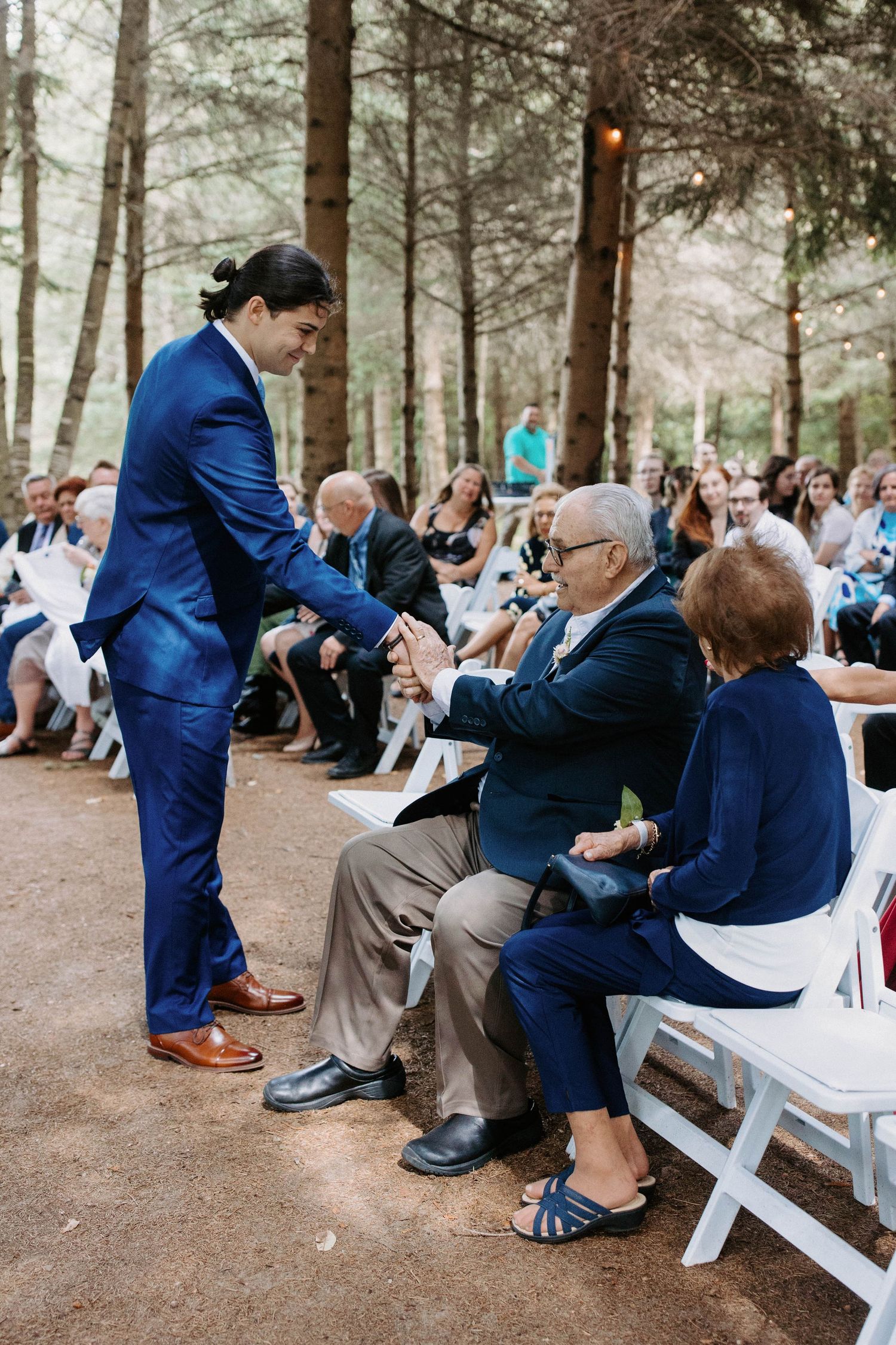 Wedding guests seated on white chairs in a pine forest setting during an outdoor ceremony.