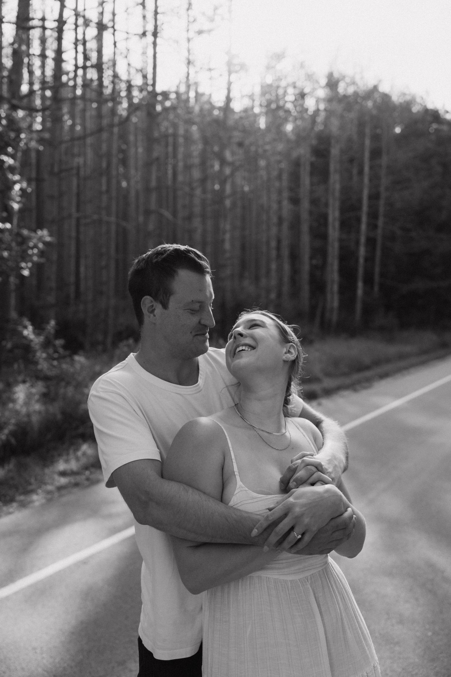A romantic couple shares playful moments together on a tree-lined road in black and white.