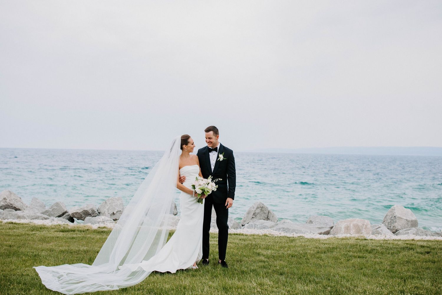 Wedding couple shares romantic moment on grassy cliff by turquoise ocean waters.