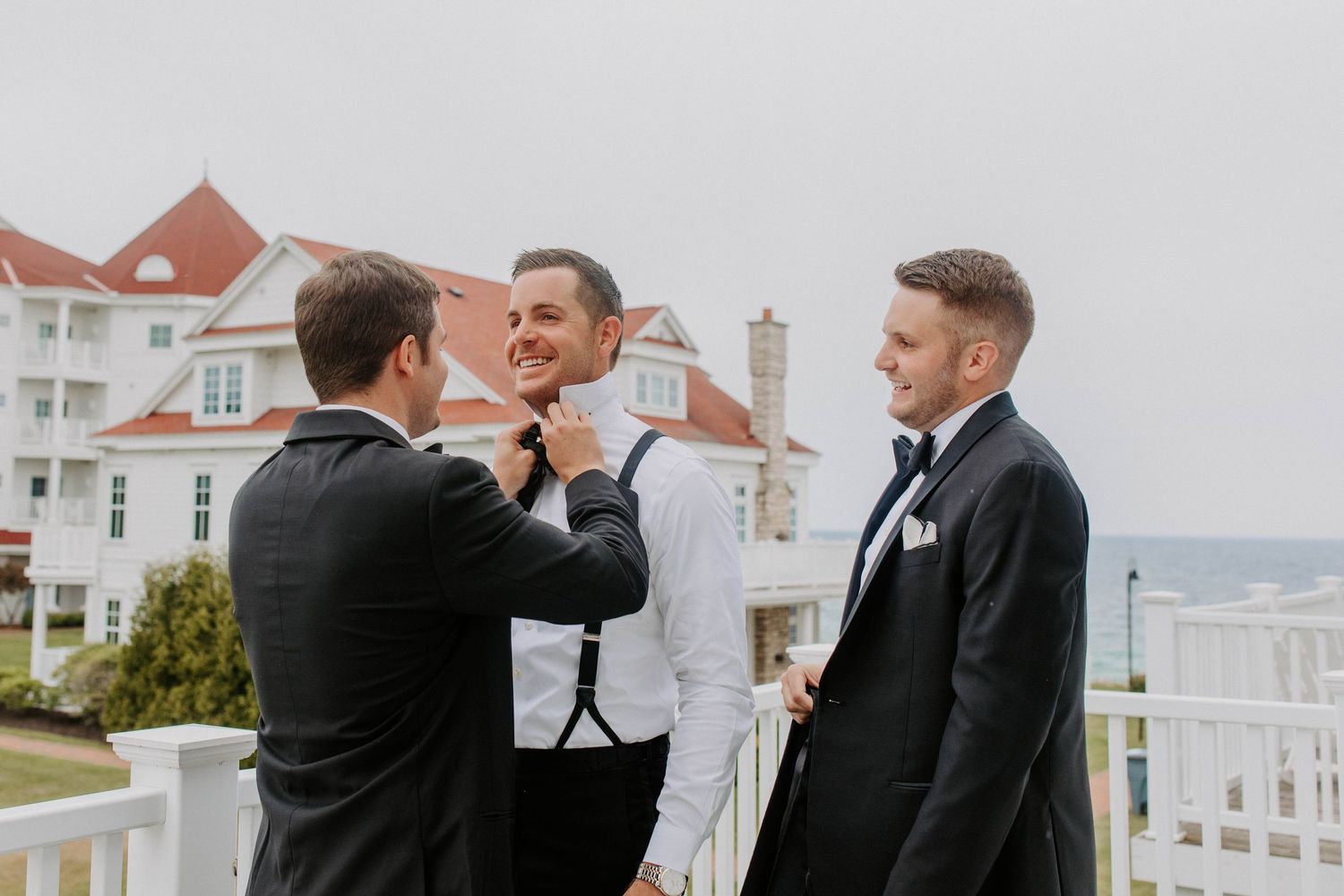 Wedding party sharing laughs on coastal property with white houses.