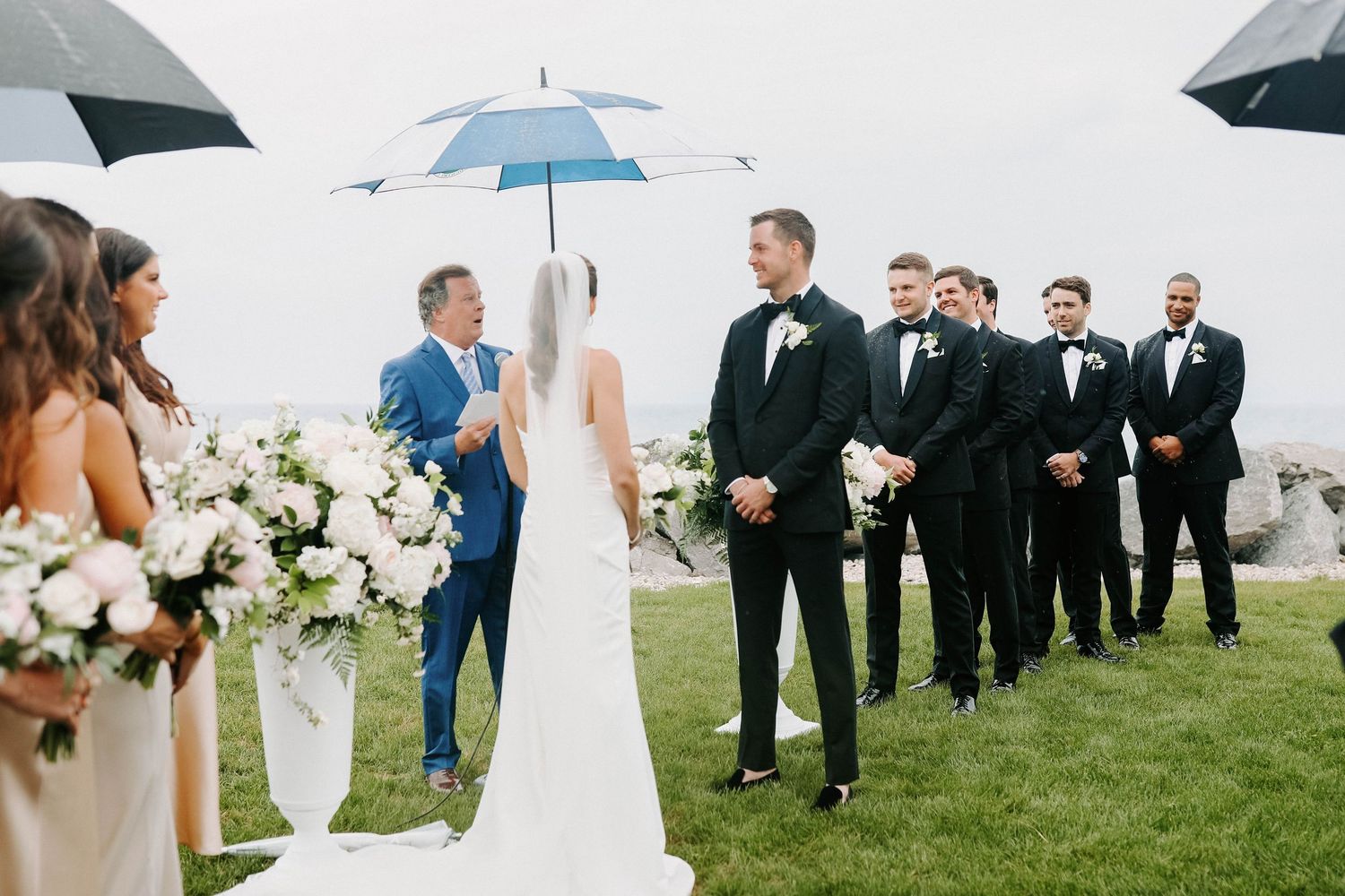 Wedding party holding white umbrellas poses together on a cloudy day outdoors.