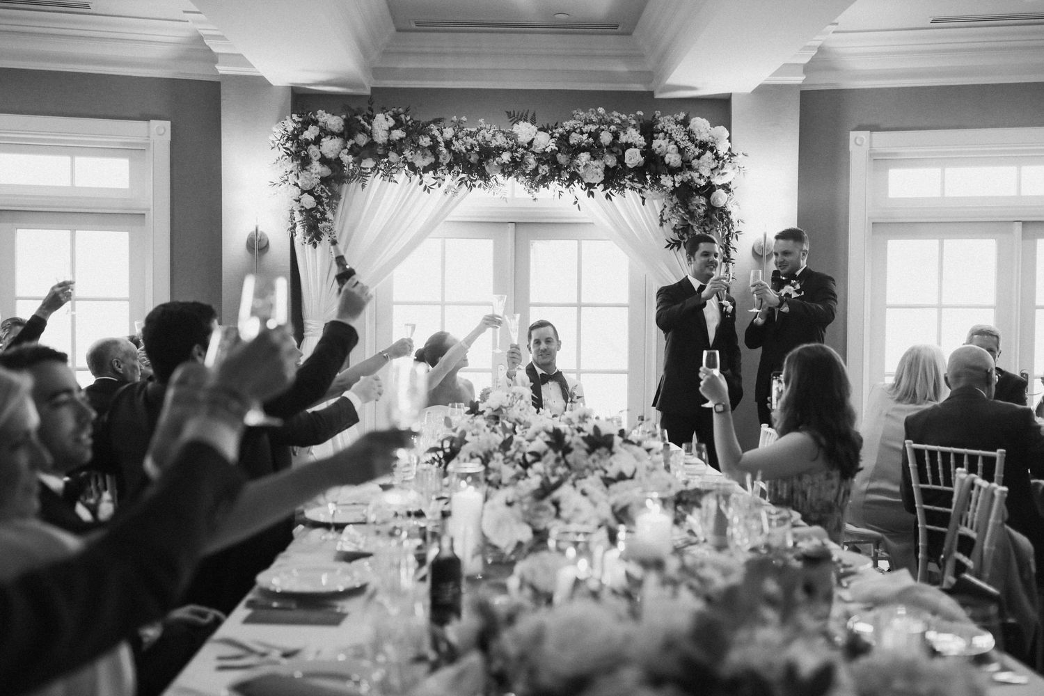 Wedding reception hall with elegant floral decor and guests celebrating at decorated tables.