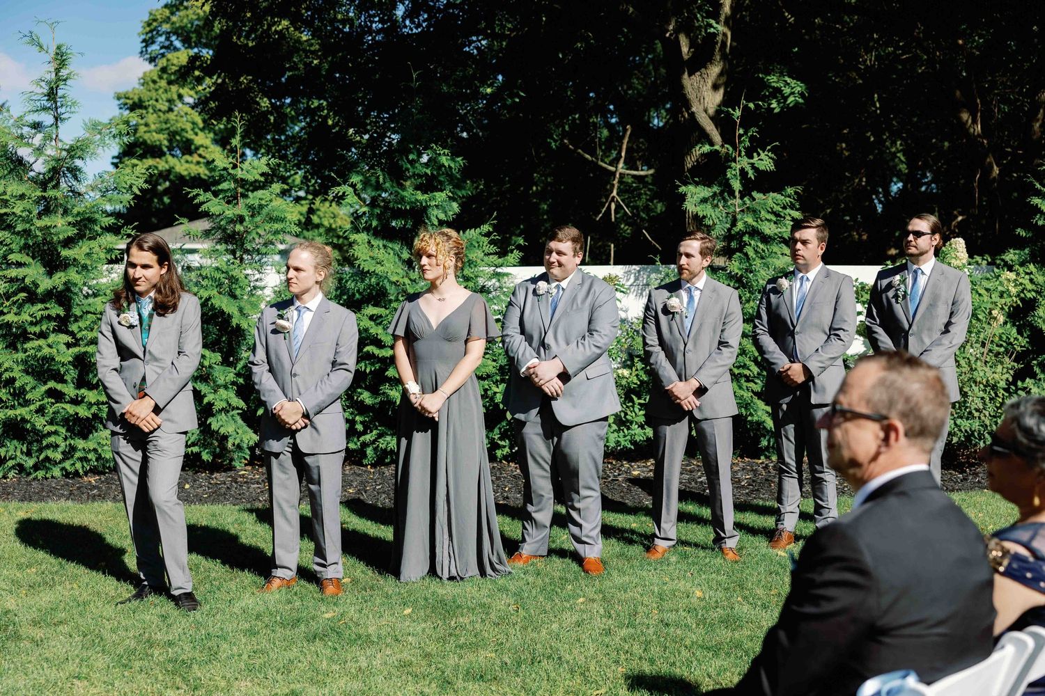 Wedding party members in gray suits stand together on a sunny lawn during an outdoor ceremony.