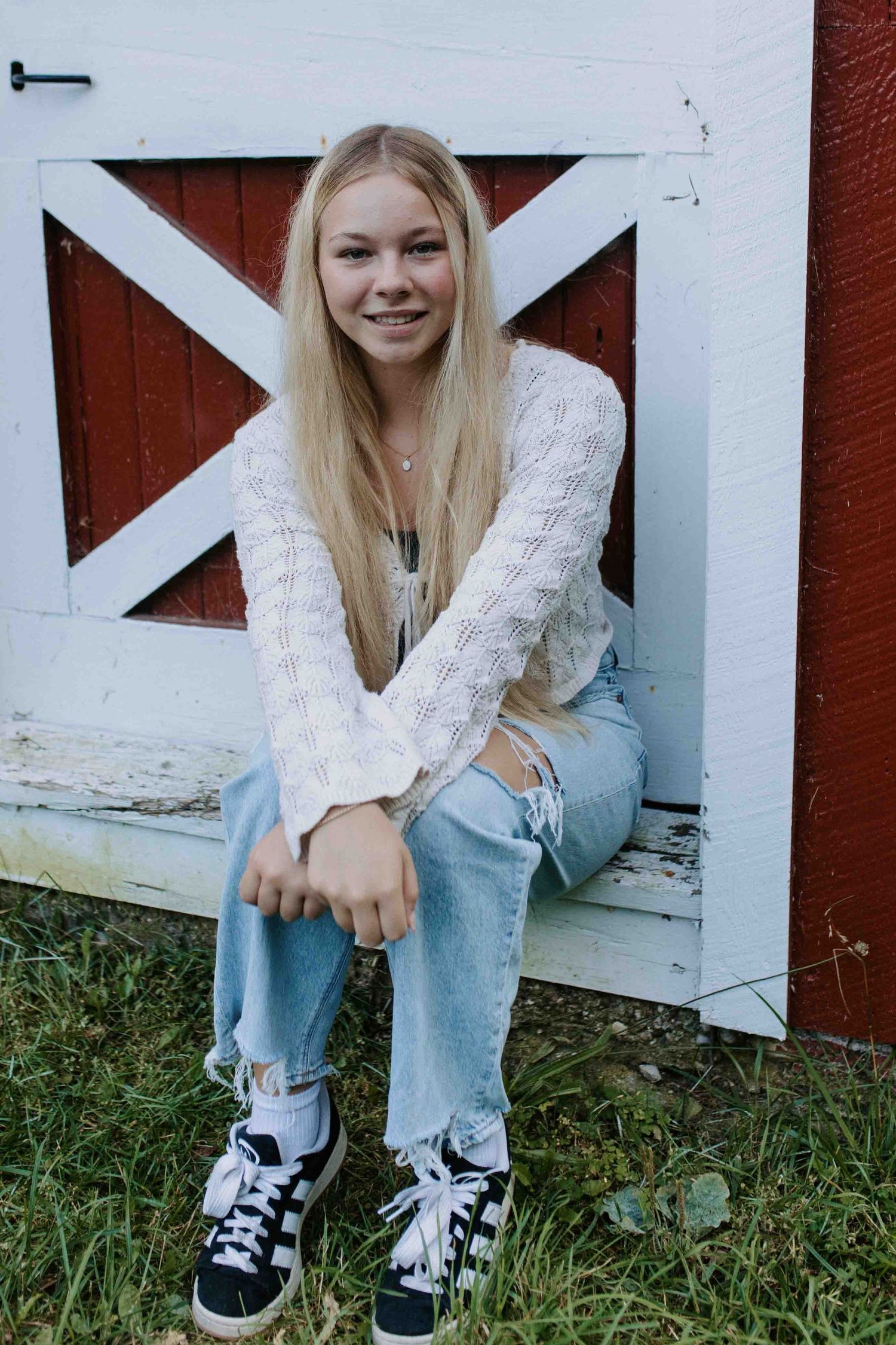 Person in ripped jeans and white sweater sits against white fence with red barn in background.