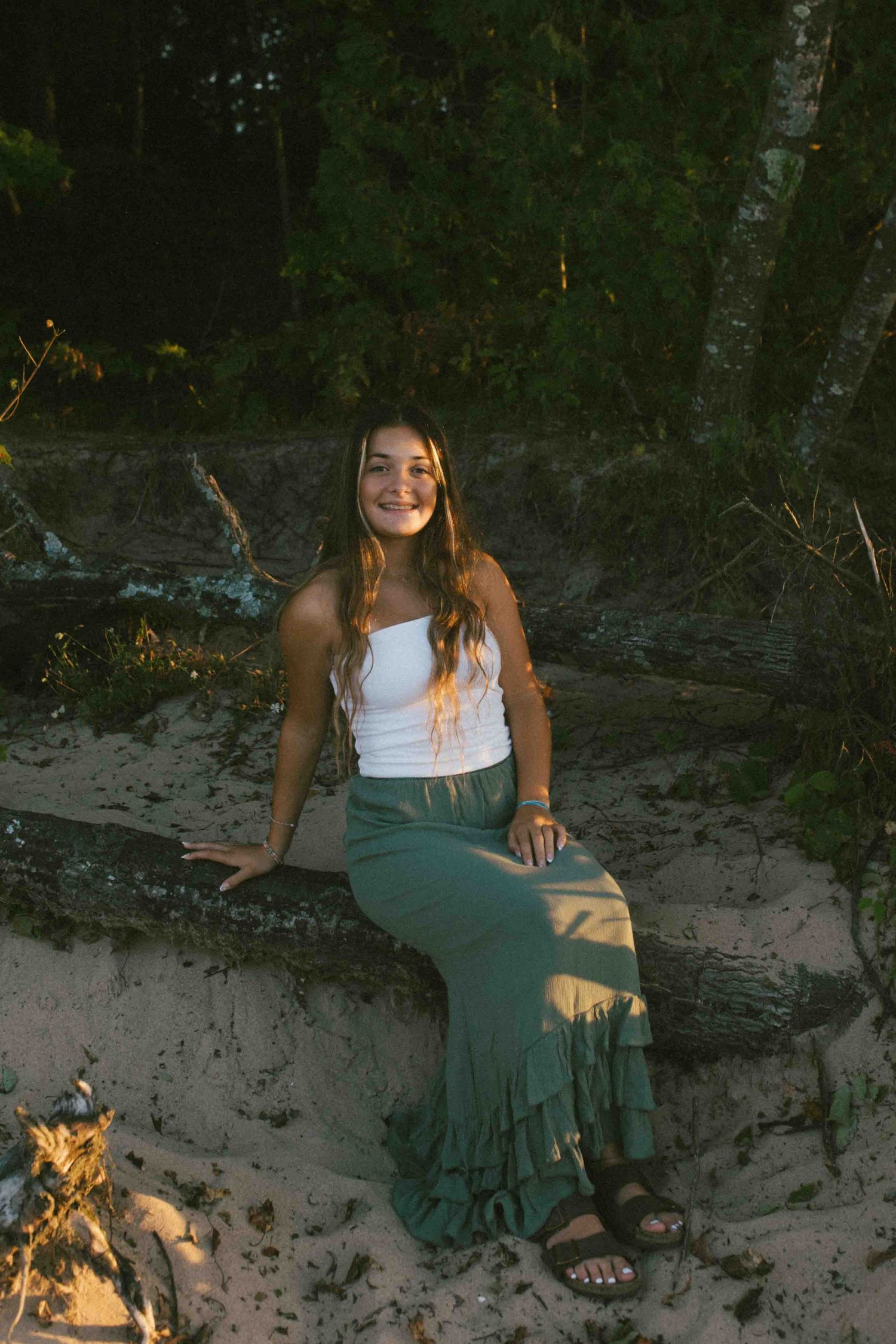 A person in casual summer attire sits on a sandy embankment with trees in the background during golden hour.