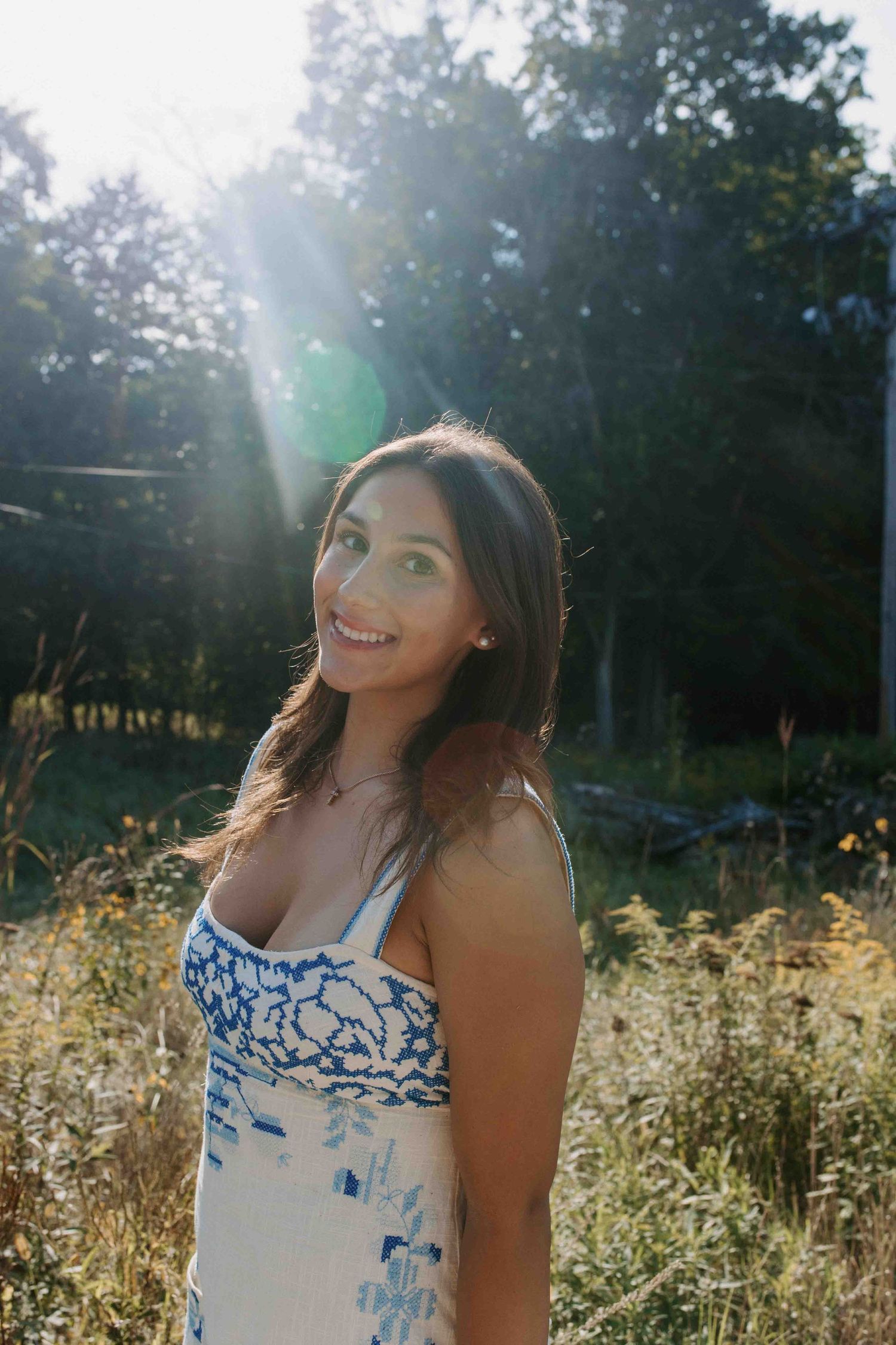 A floral sundress-clad figure poses playfully in a sunlit meadow with golden grass and trees in the background.