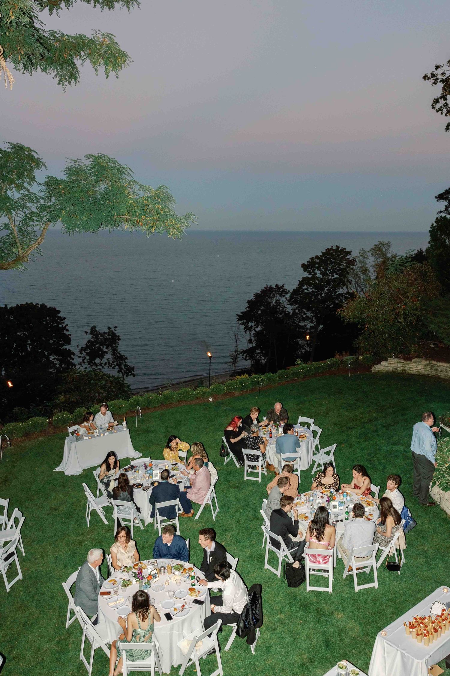 Outdoor wedding reception on a lawn overlooking the ocean with guests seated at round tables at dusk.