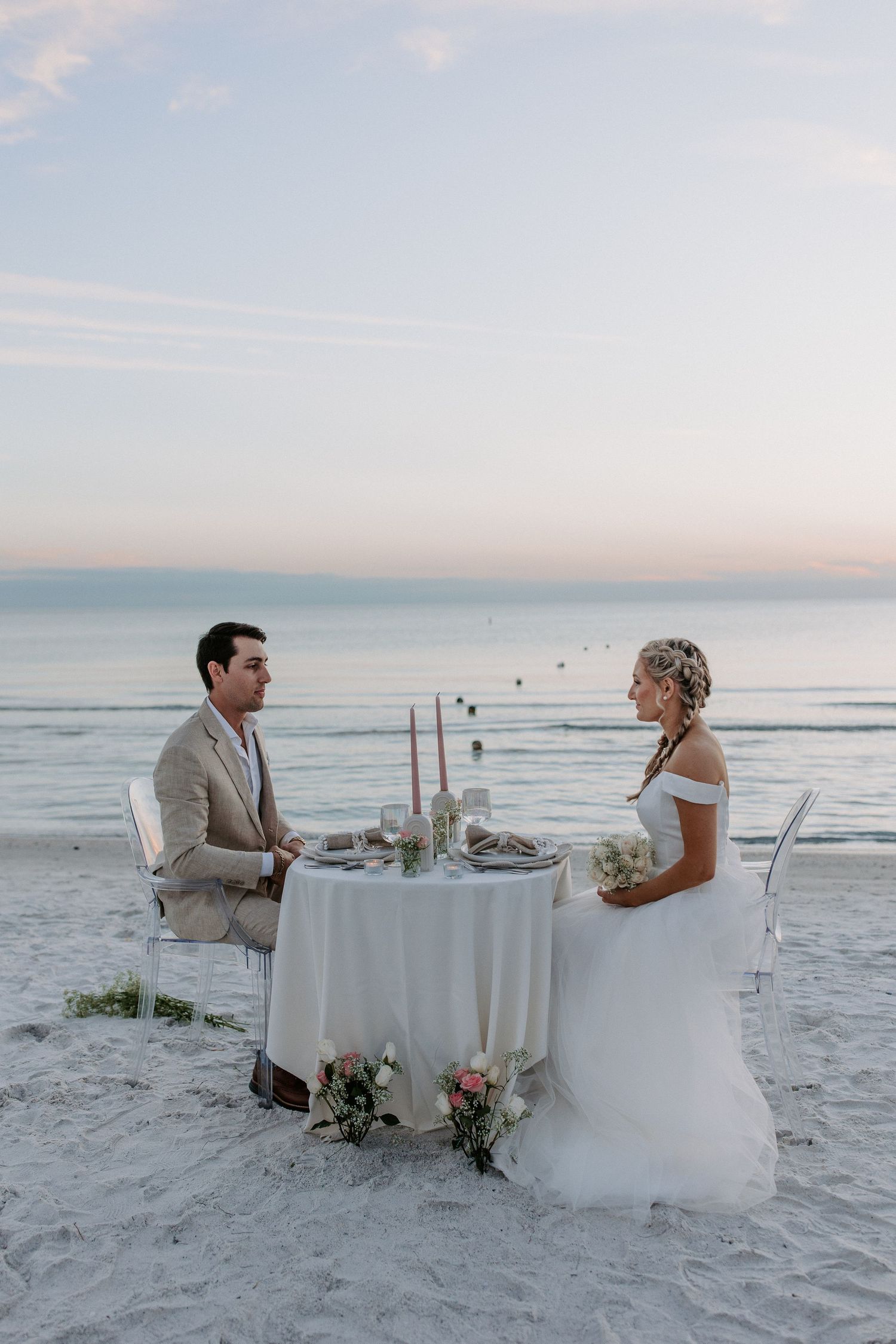 Elegant beachside dinner setup with white tablecloths and intimate seating on pristine sand at dusk.