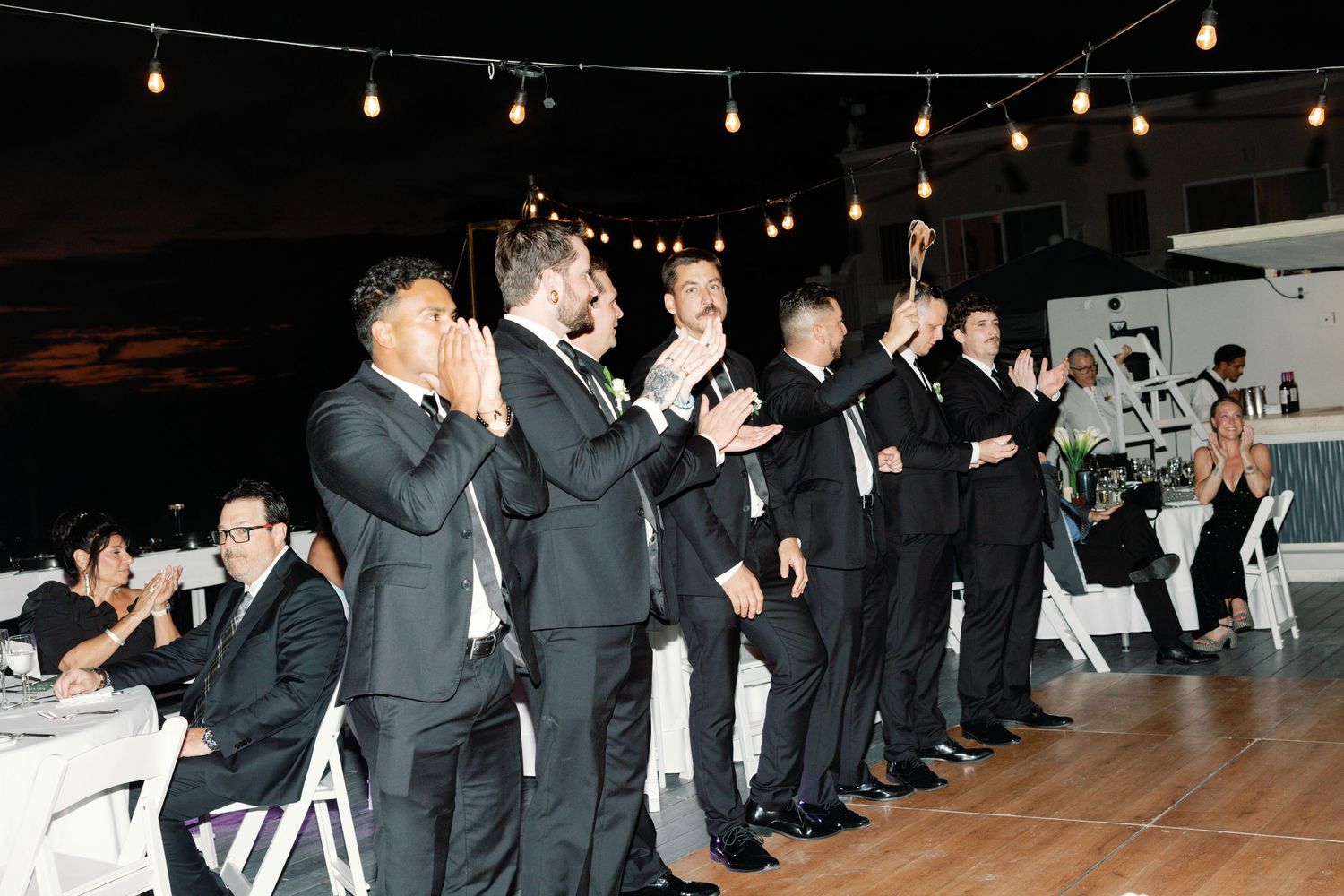 Groomsmen in black suits cheer and celebrate during a wedding reception under string lights.