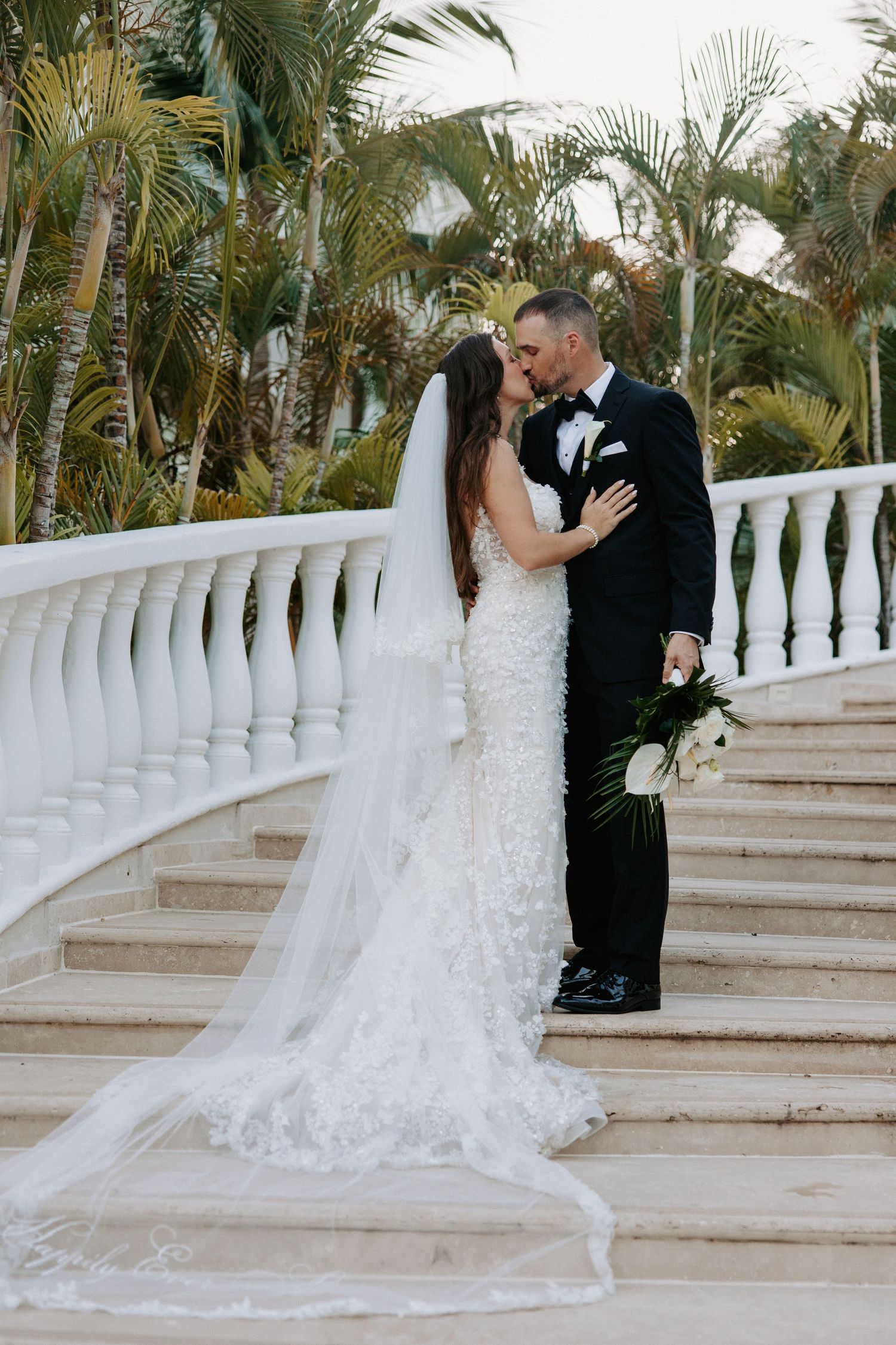 Romantic couple poses on white stone staircase surrounded by palm trees.