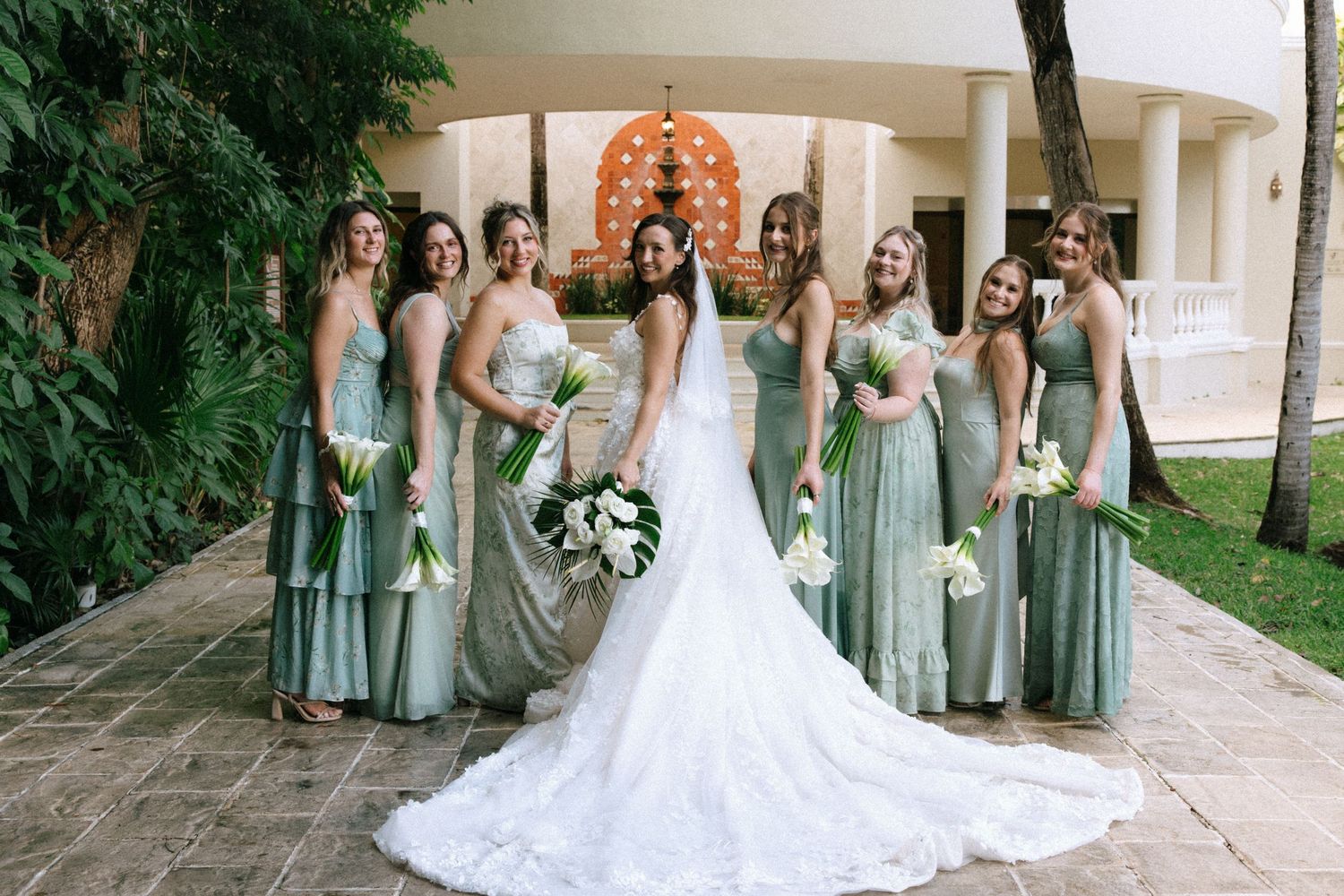 Wedding party in mint green dresses gathered around bride in flowing white gown on stone patio.