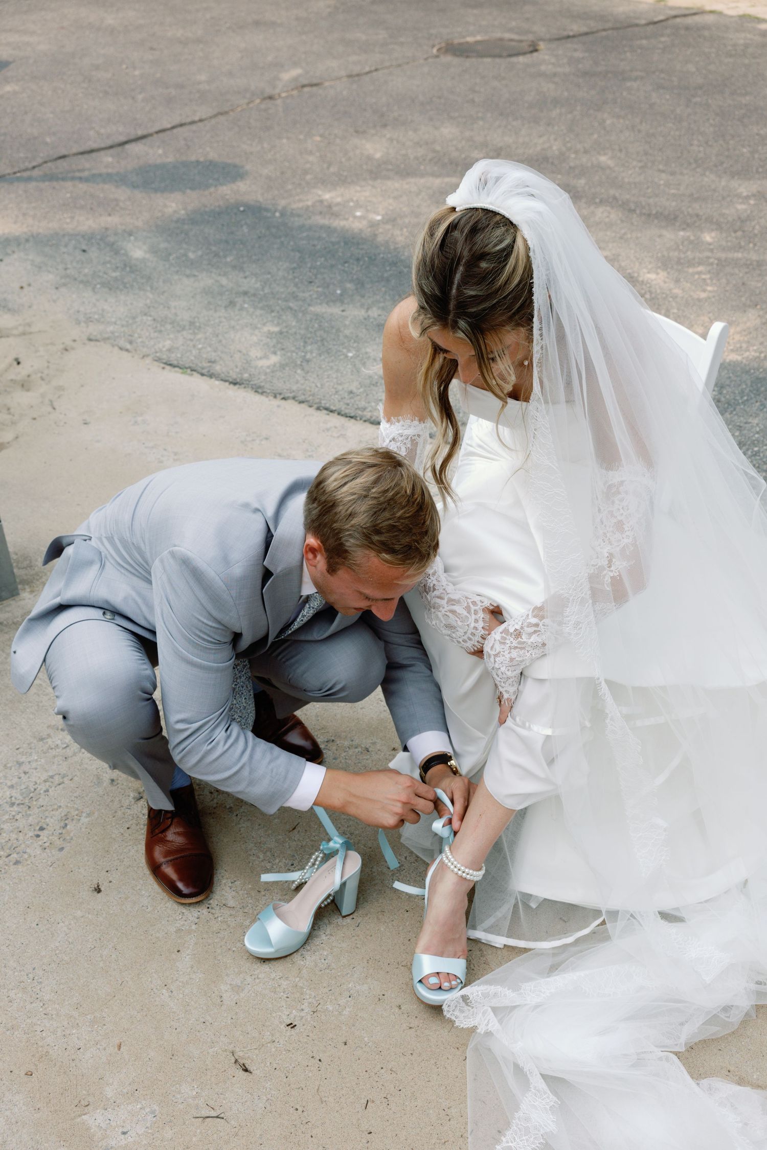 Someone in a light gray suit helps adjust a white wedding dress on a concrete surface.