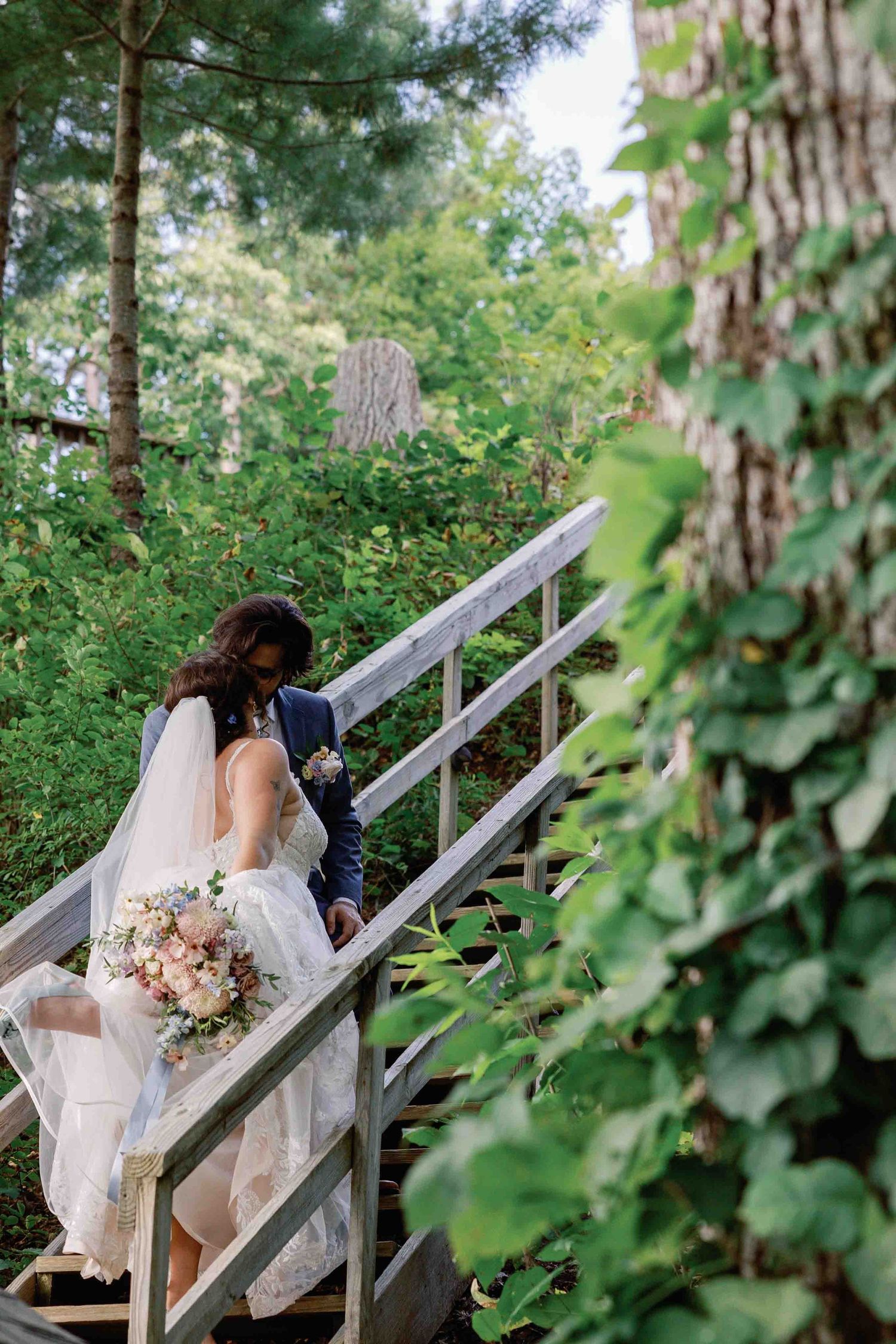 Wedding couple poses on rustic wooden stairs surrounded by lush summer foliage.