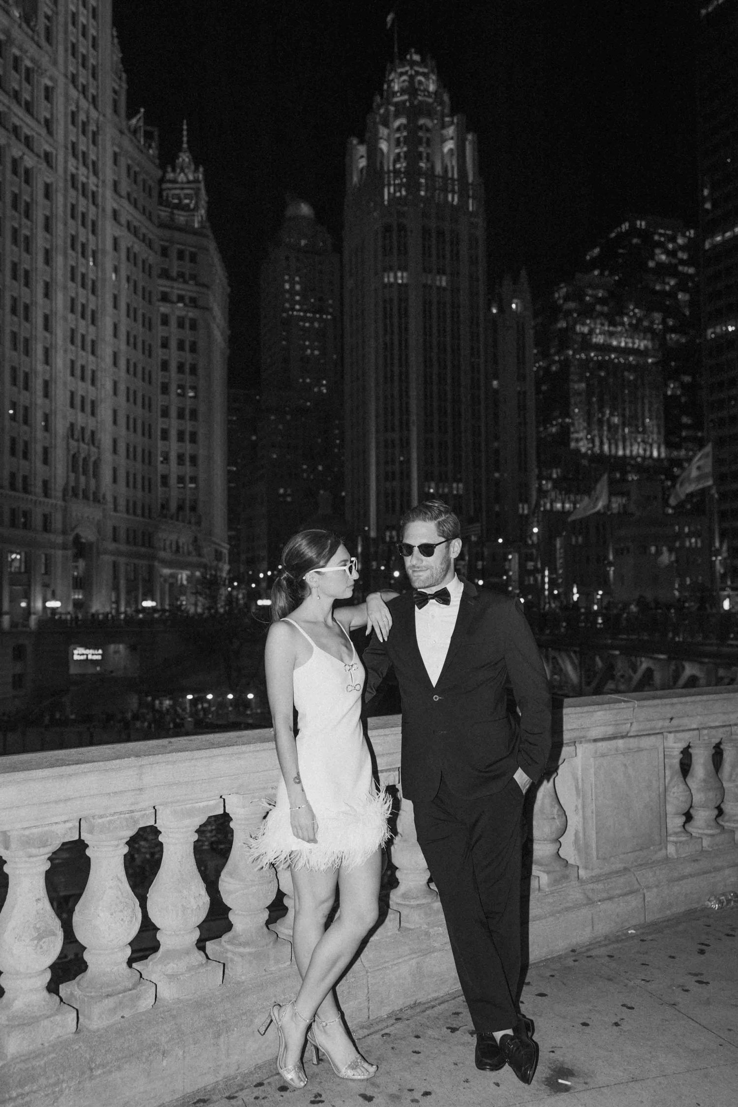 A couple in formal attire poses romantically along an illuminated city bridge at night.