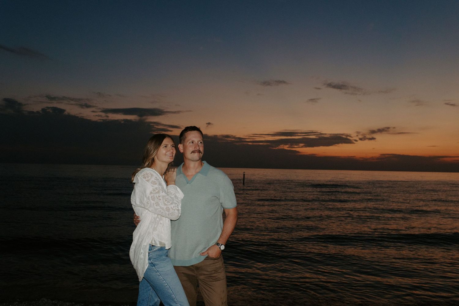 A couple embracing on the beach during a dramatic sunset with waves in the background.