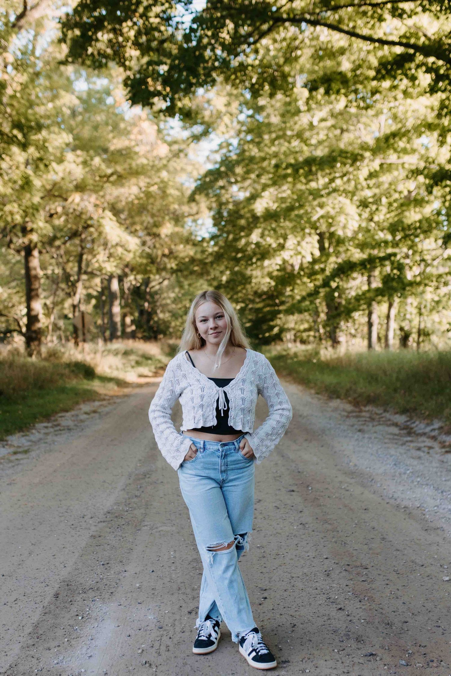 Person in denim and lace poses on dirt path under sunlit trees.