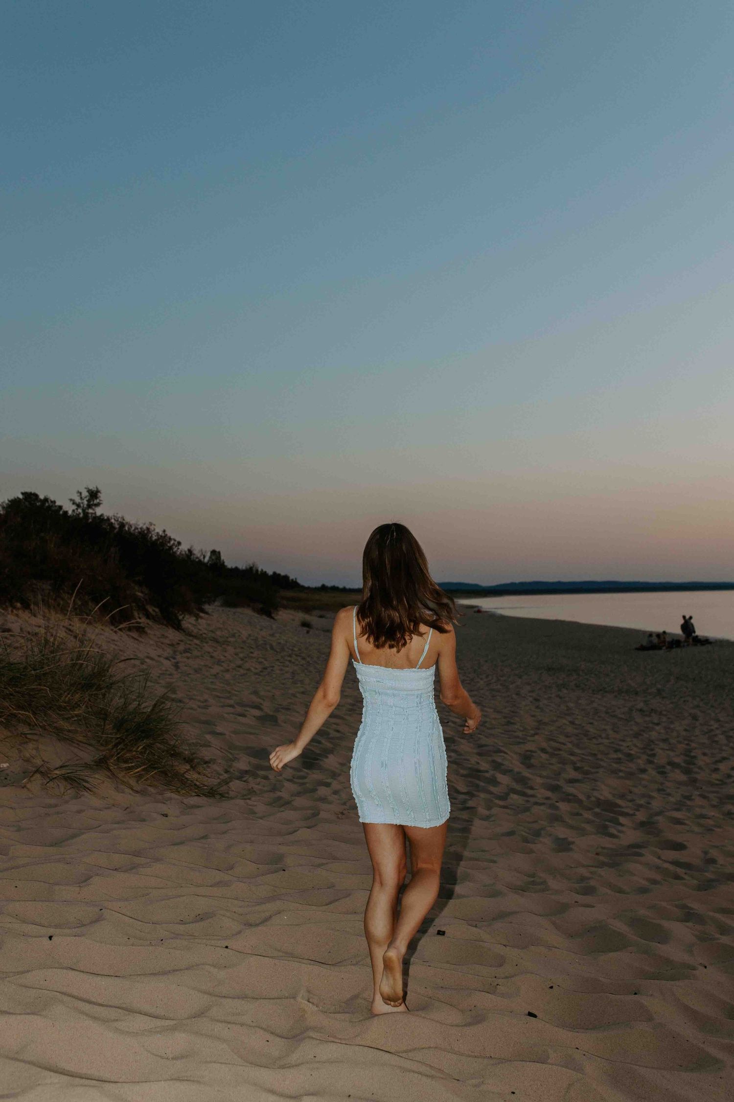 A light blue dress silhouette walks along sandy dunes at sunset against a tranquil ocean backdrop.
