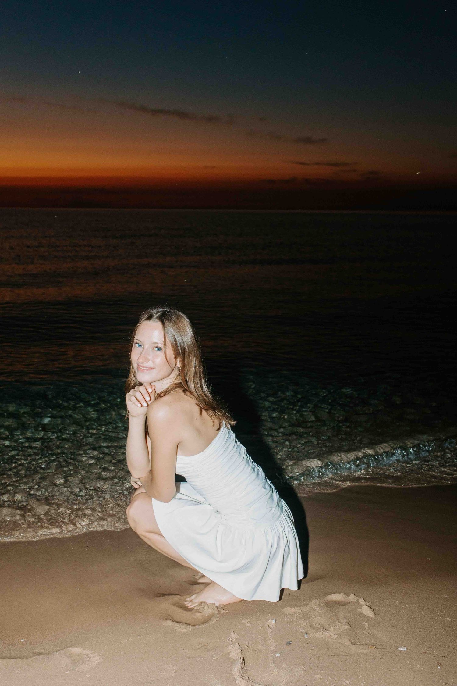 A person in white lounges on the sandy beach during a vibrant sunset over the ocean.