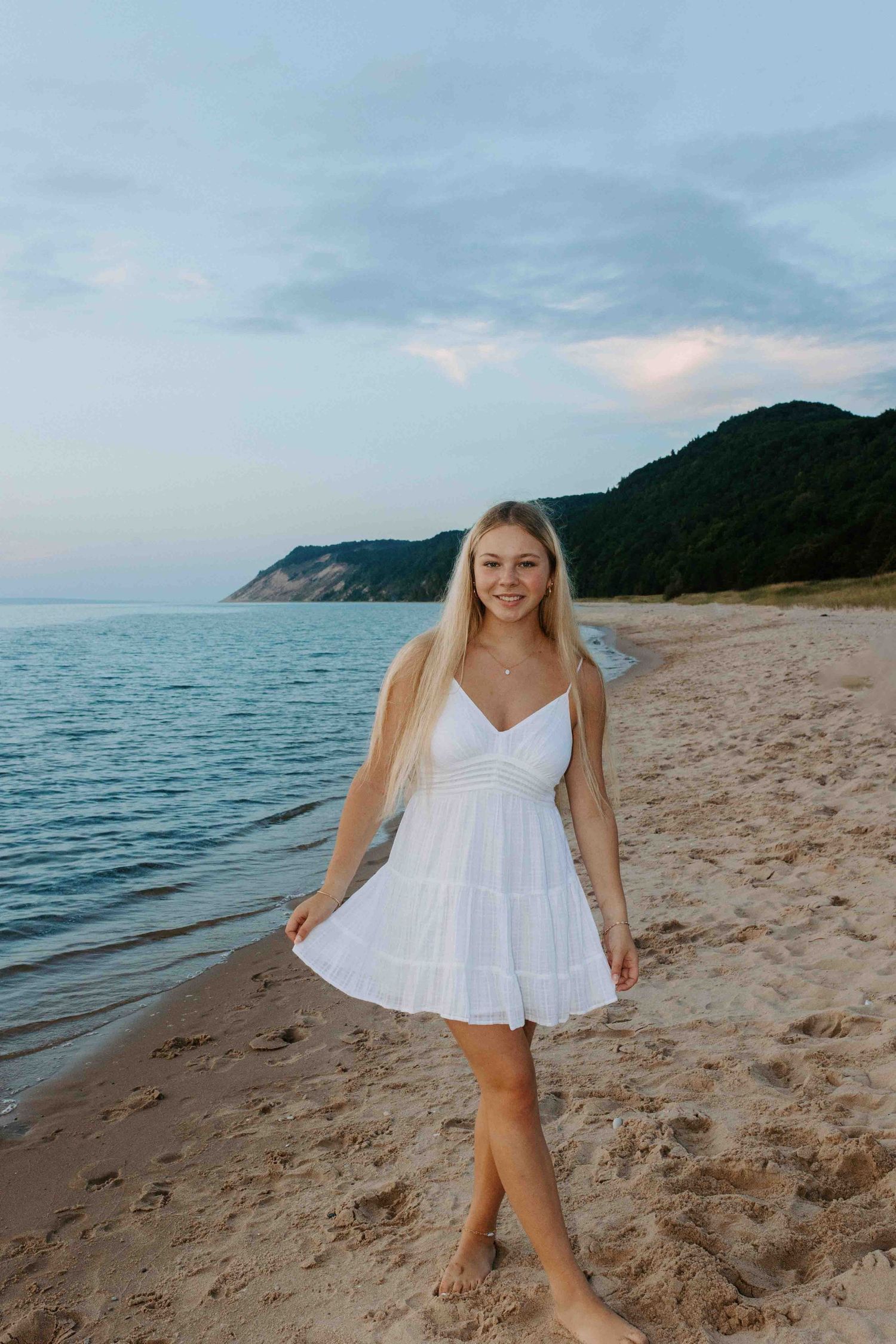 Candid beach photoshoot with someone in a flowy white dress walking along the sandy shoreline.