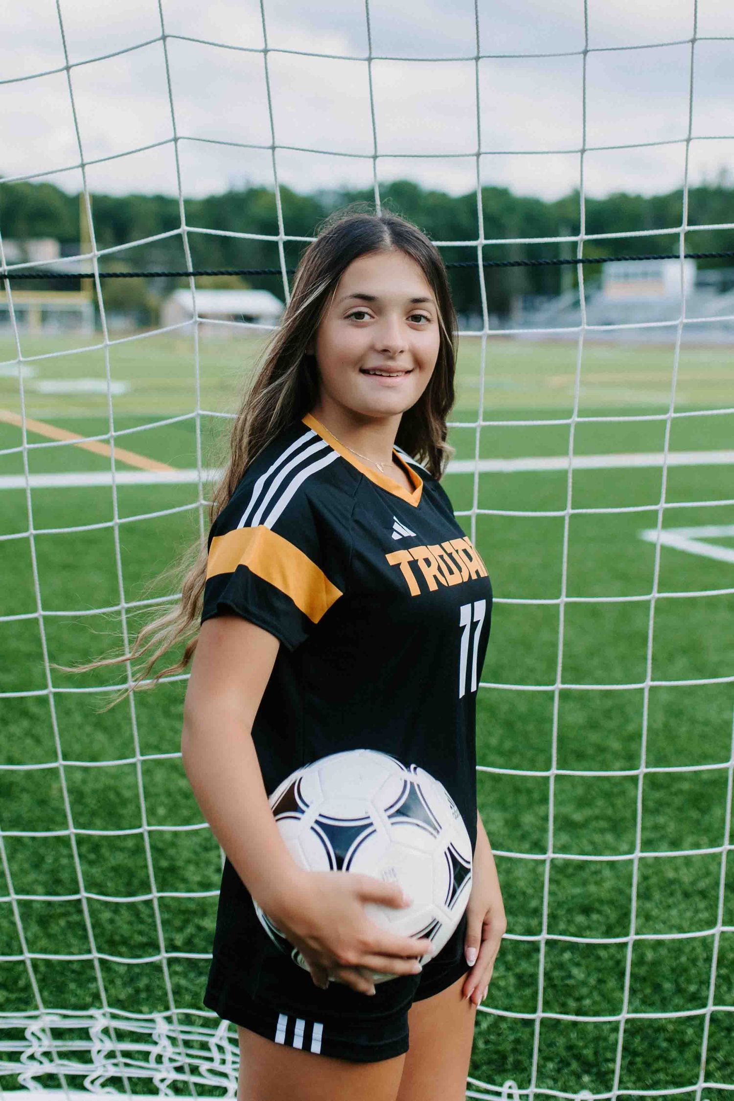 Soccer player wearing black and gold uniform poses with ball in front of goal net.