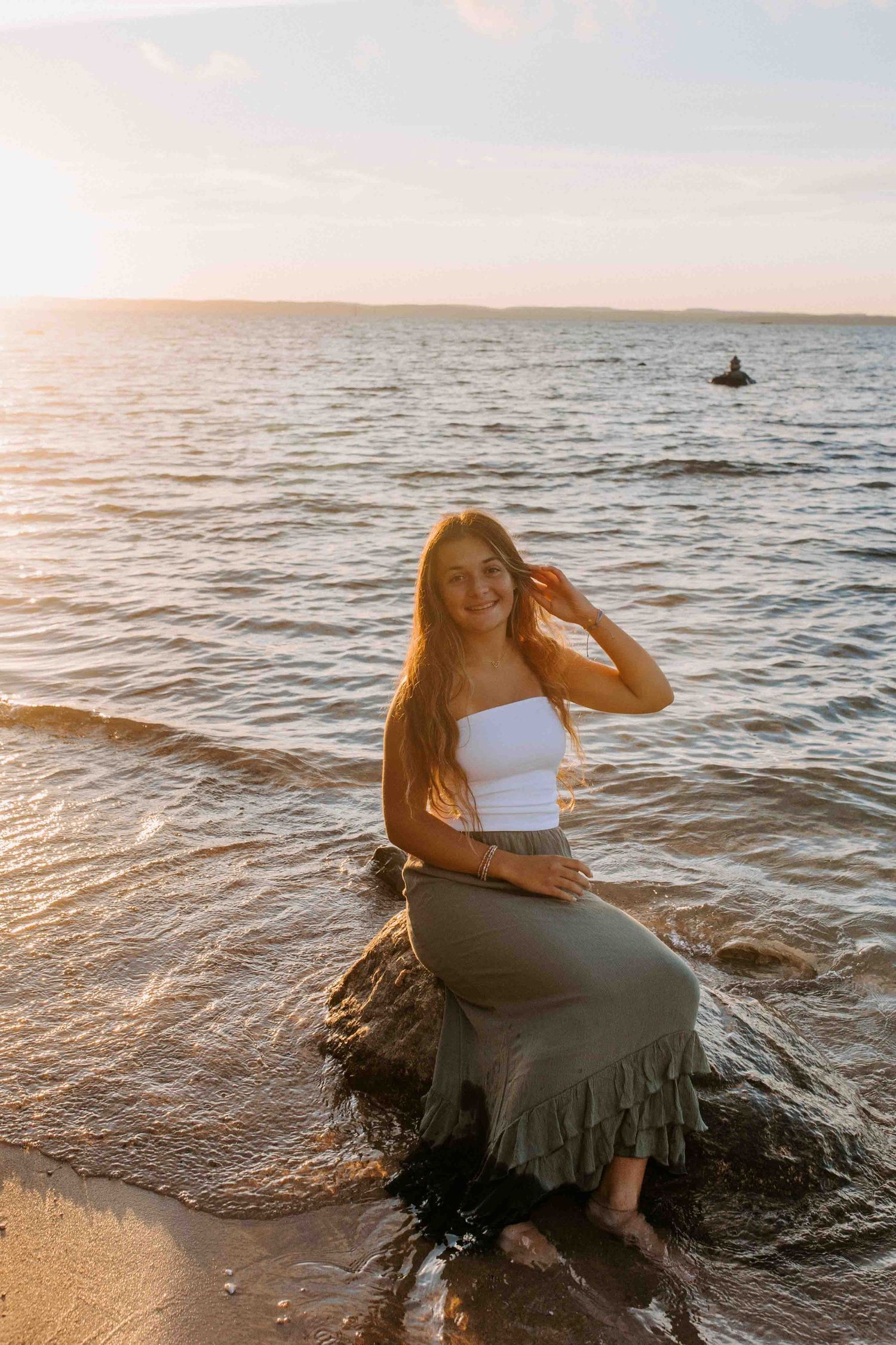 A person in beachwear sits on coastal rocks during golden hour with waves in the background.