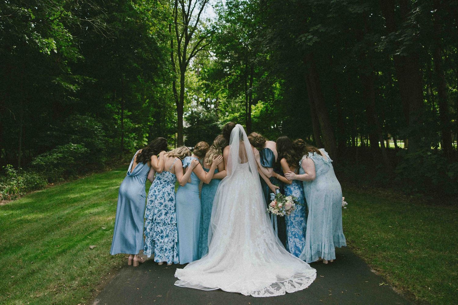Wedding party poses outdoors in a wooded area with bridesmaids in light blue dresses and groomsmen in dark suits.
