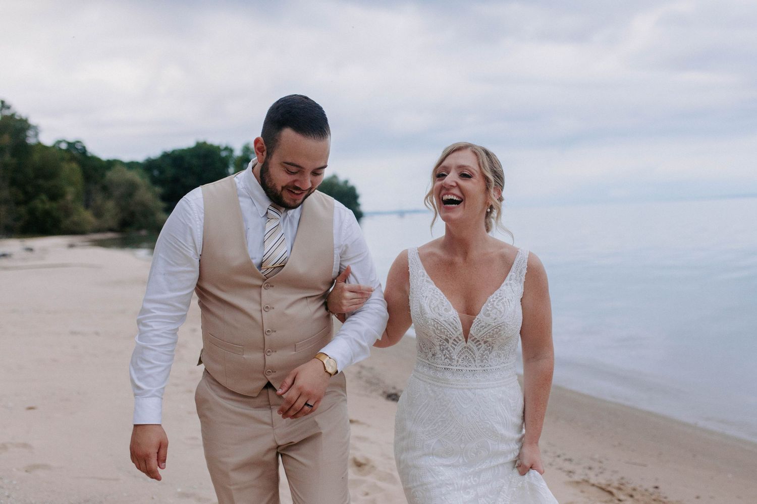 Wedding party laughs and celebrates together on a sandy beach with ocean waves in the background.