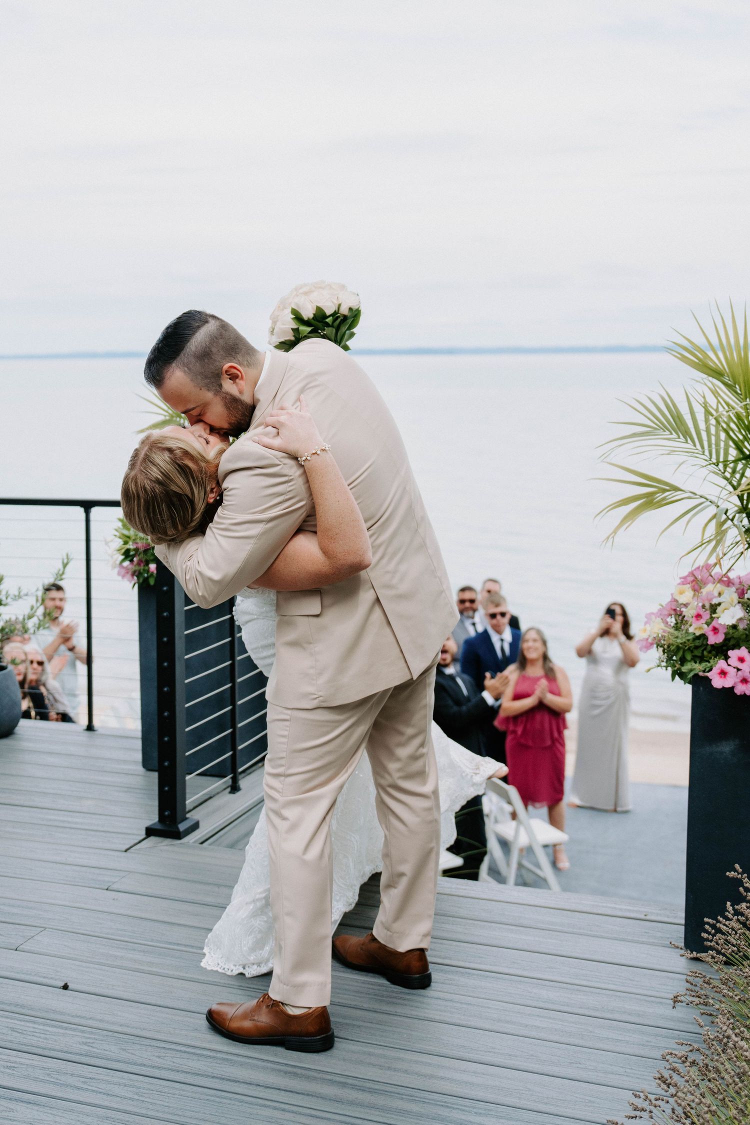 Romantic dip kiss during a waterfront wedding ceremony with guests watching in the background.