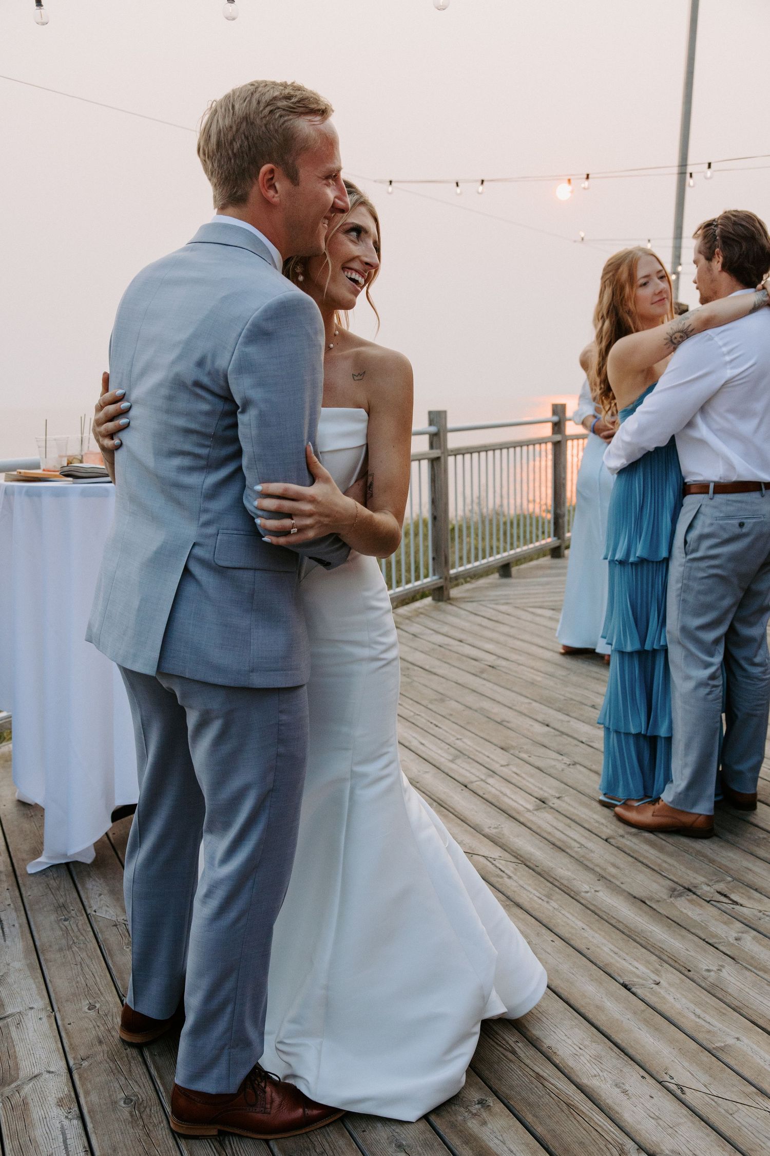 Wedding couple hugs on a rustic wooden deck with guests and white decor visible in the background.