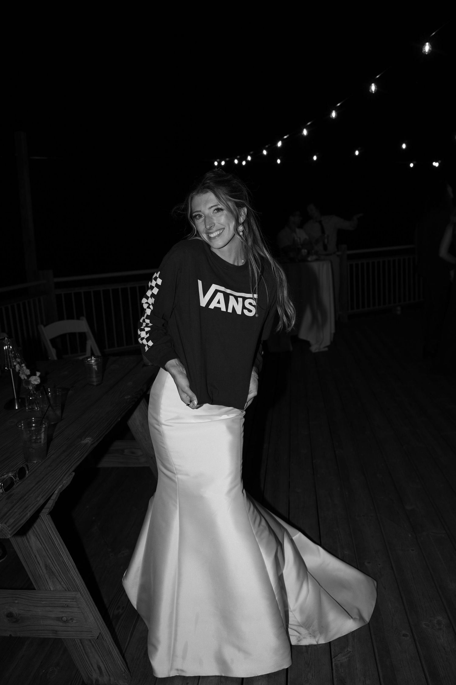A person in a red Vans sweatshirt and white dress spins on a wooden deck at night under string lights.