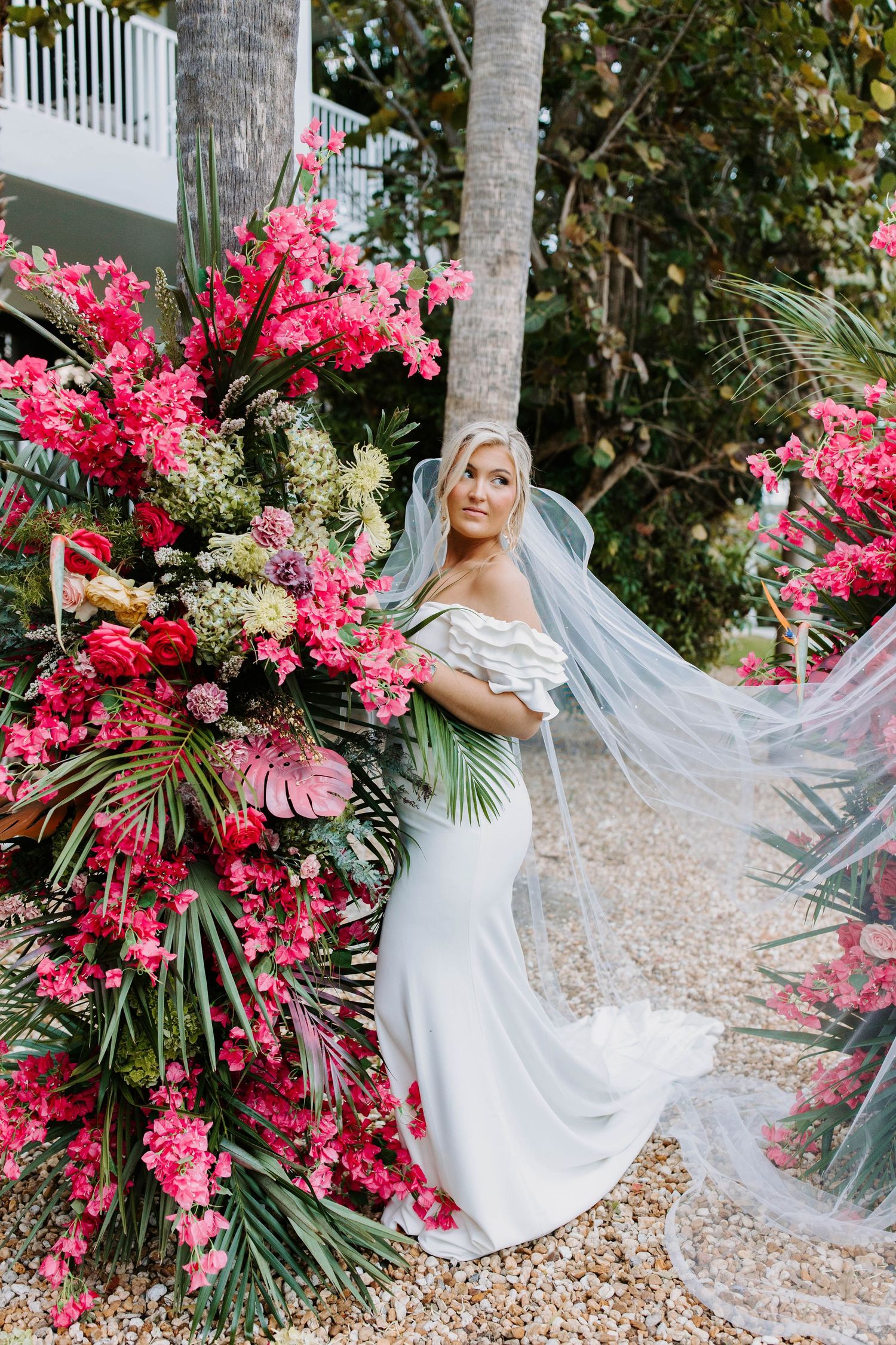 Bride in flowing white gown poses dramatically with pink bougainvillea flowers and palm trees.