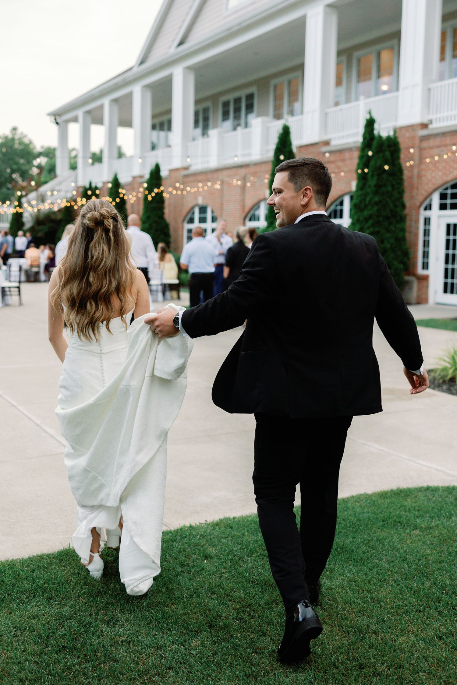 Wedding couple walking playfully on grass in front of elegant white colonial building.