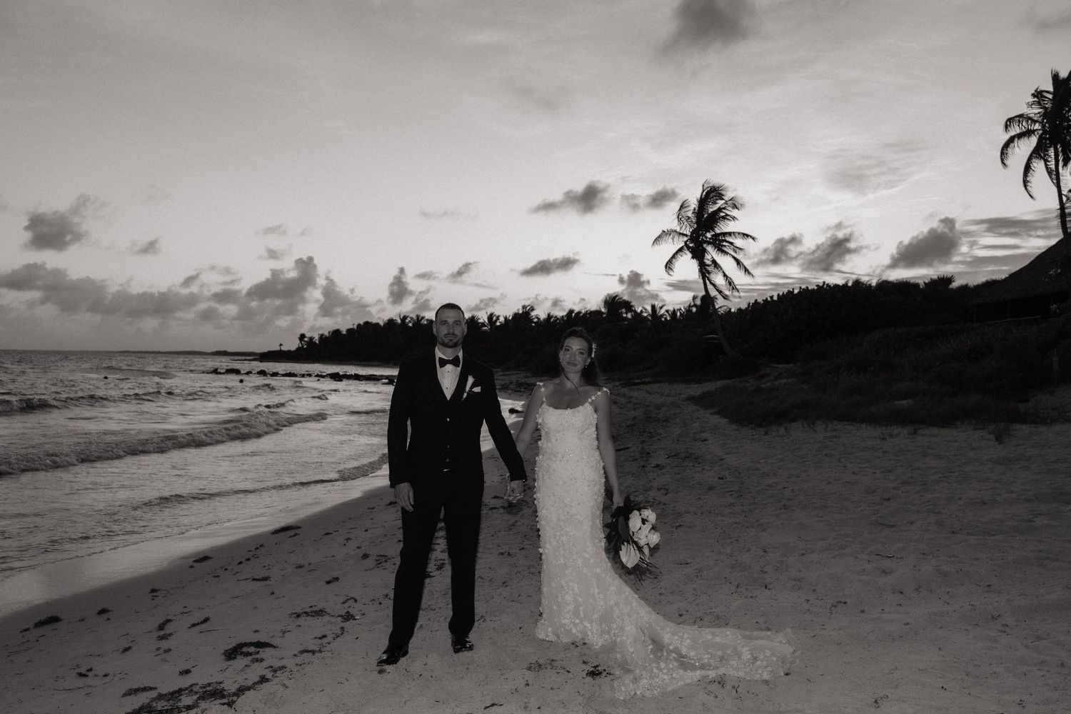 A black and white beach wedding portrait with palm trees silhouetted against cloudy sky.