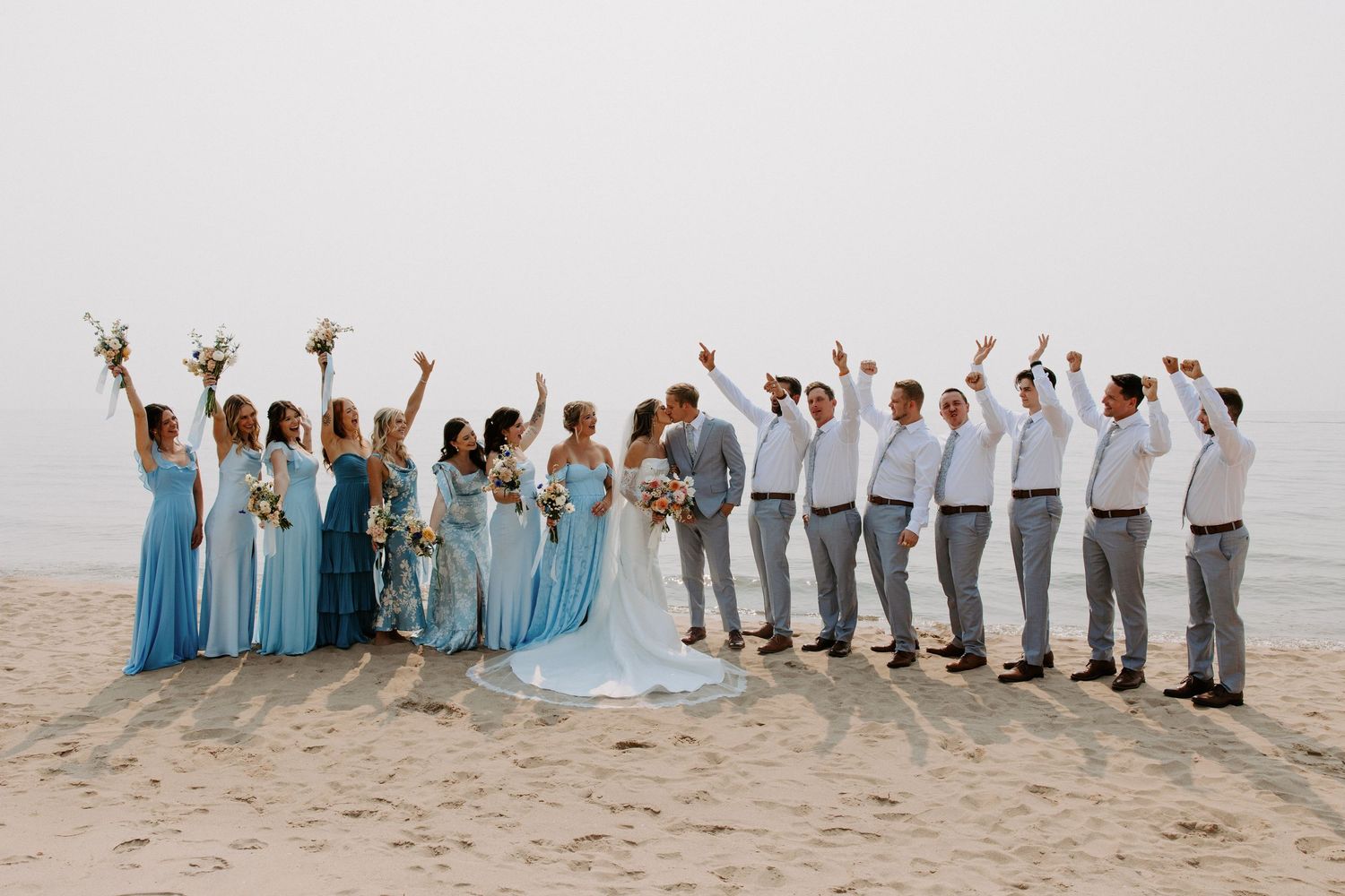 Wedding party posing together on sandy beach in coordinated light blue and gray outfits.