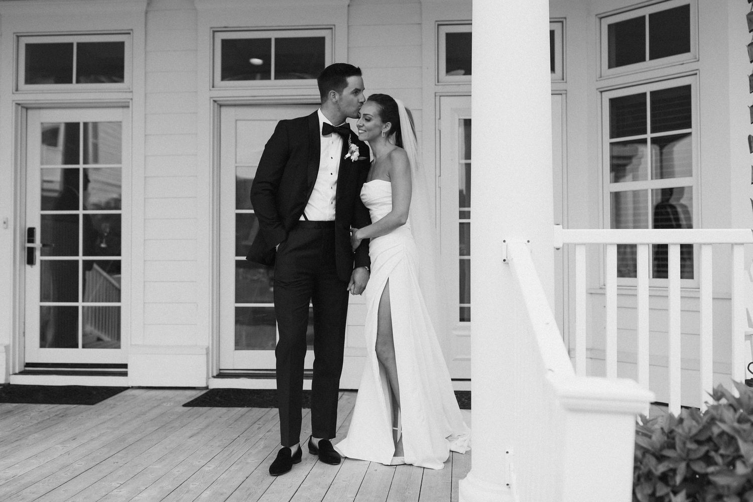 Black and white portrait of couple on white porch in formal attire.