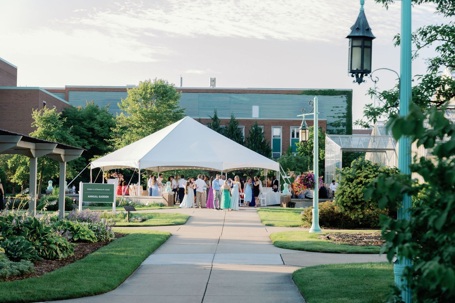 Wedding guests gather outside on a sunny day under white tents and trees for an outdoor celebration.