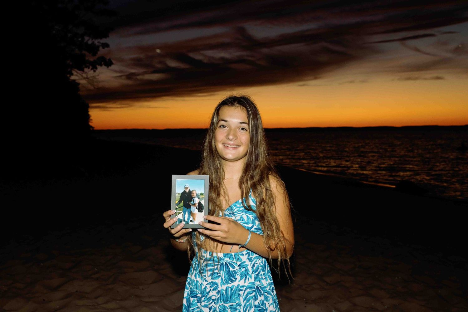 Soccer player holds a photo at sunset by the beach in a blue patterned dress.