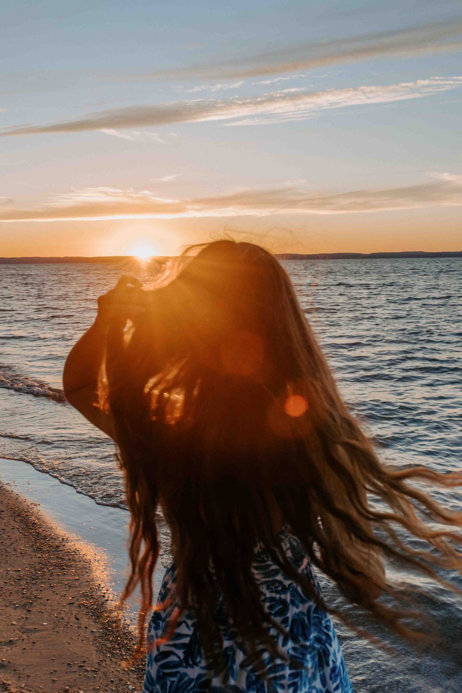 Silhouetted figure with flowing hair against orange sunset sky over ocean horizon.