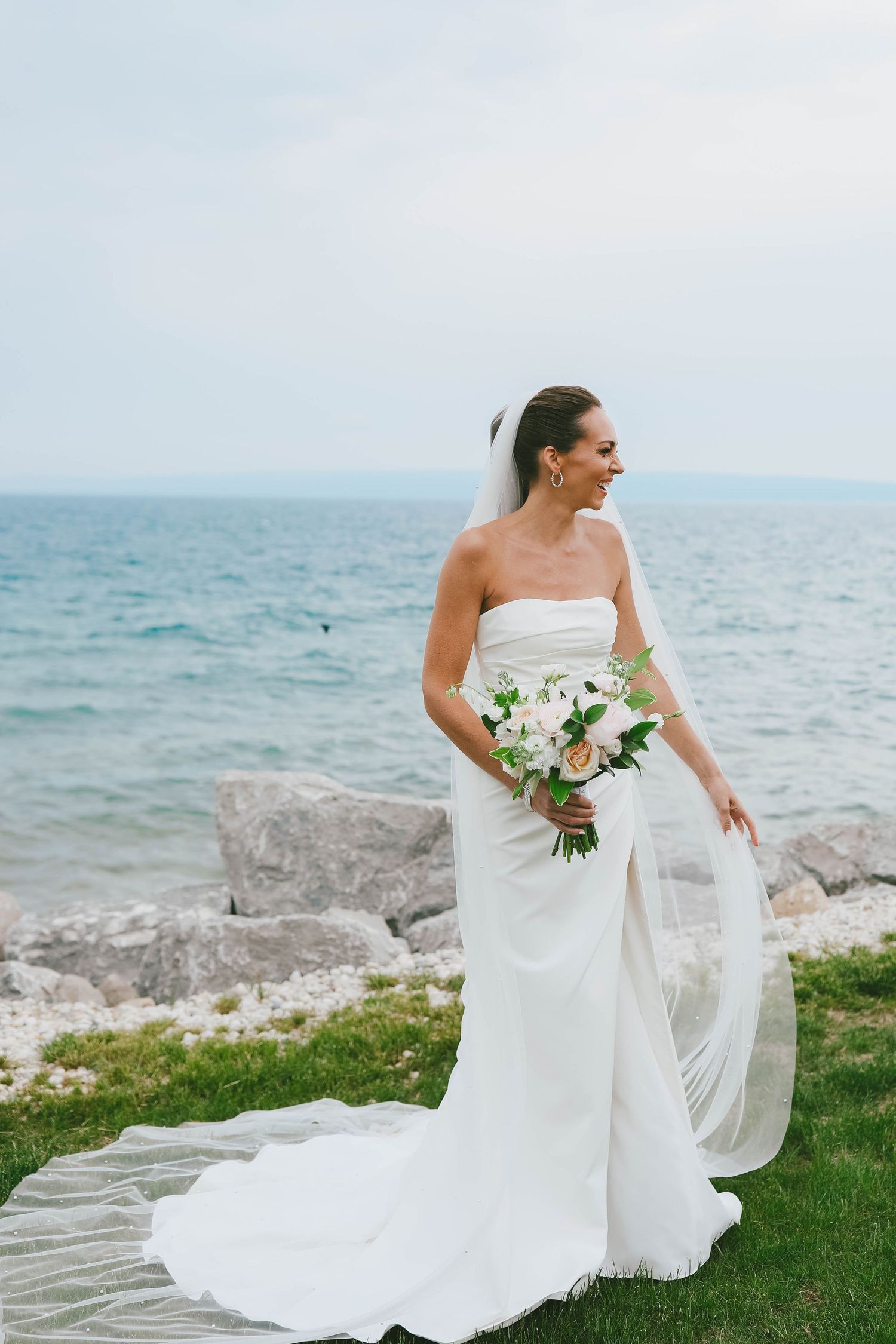 Side view of bride in flowing white gown with veil against seaside background.
