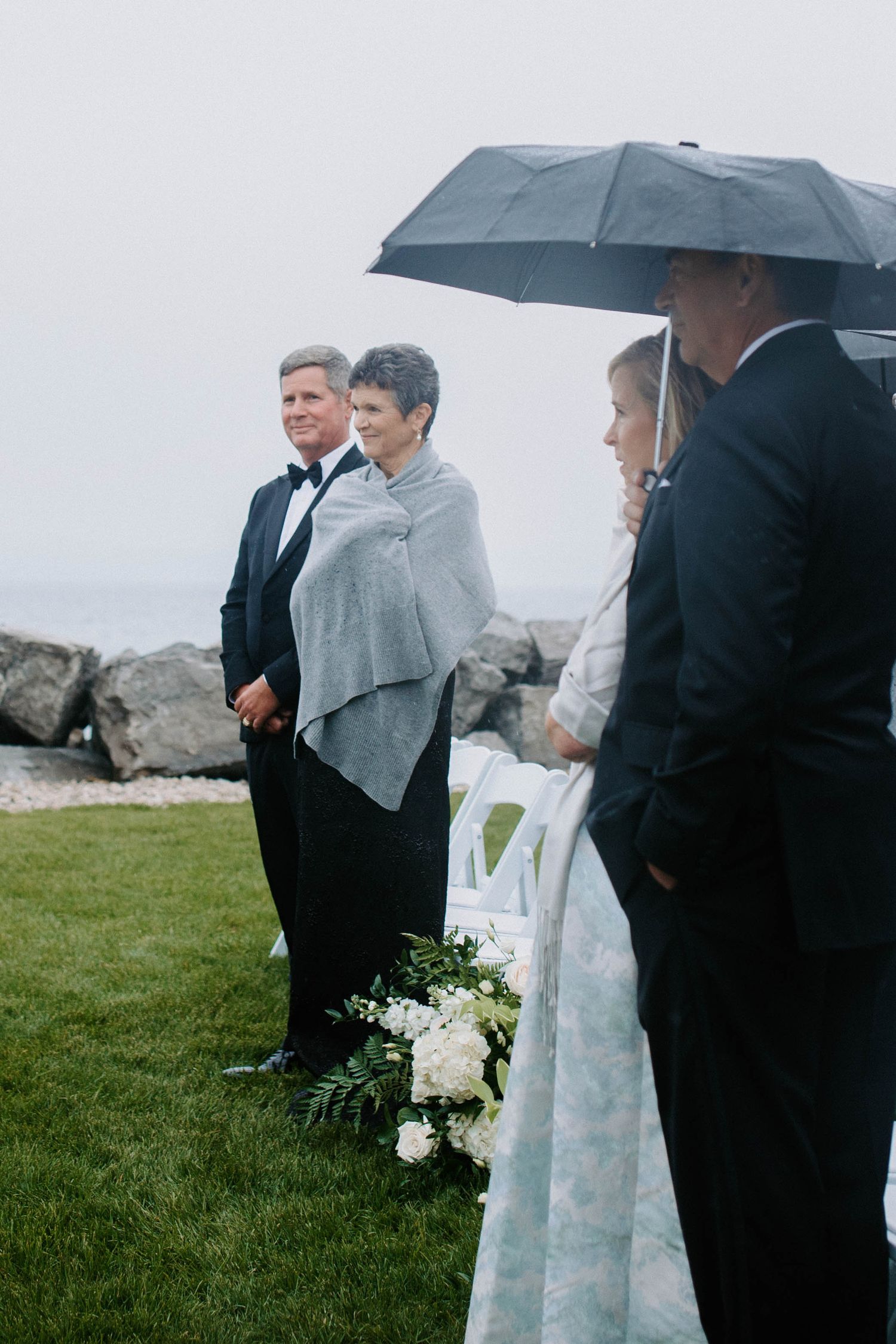 Wedding ceremony guests huddle under umbrellas on a rainy day by the ocean.