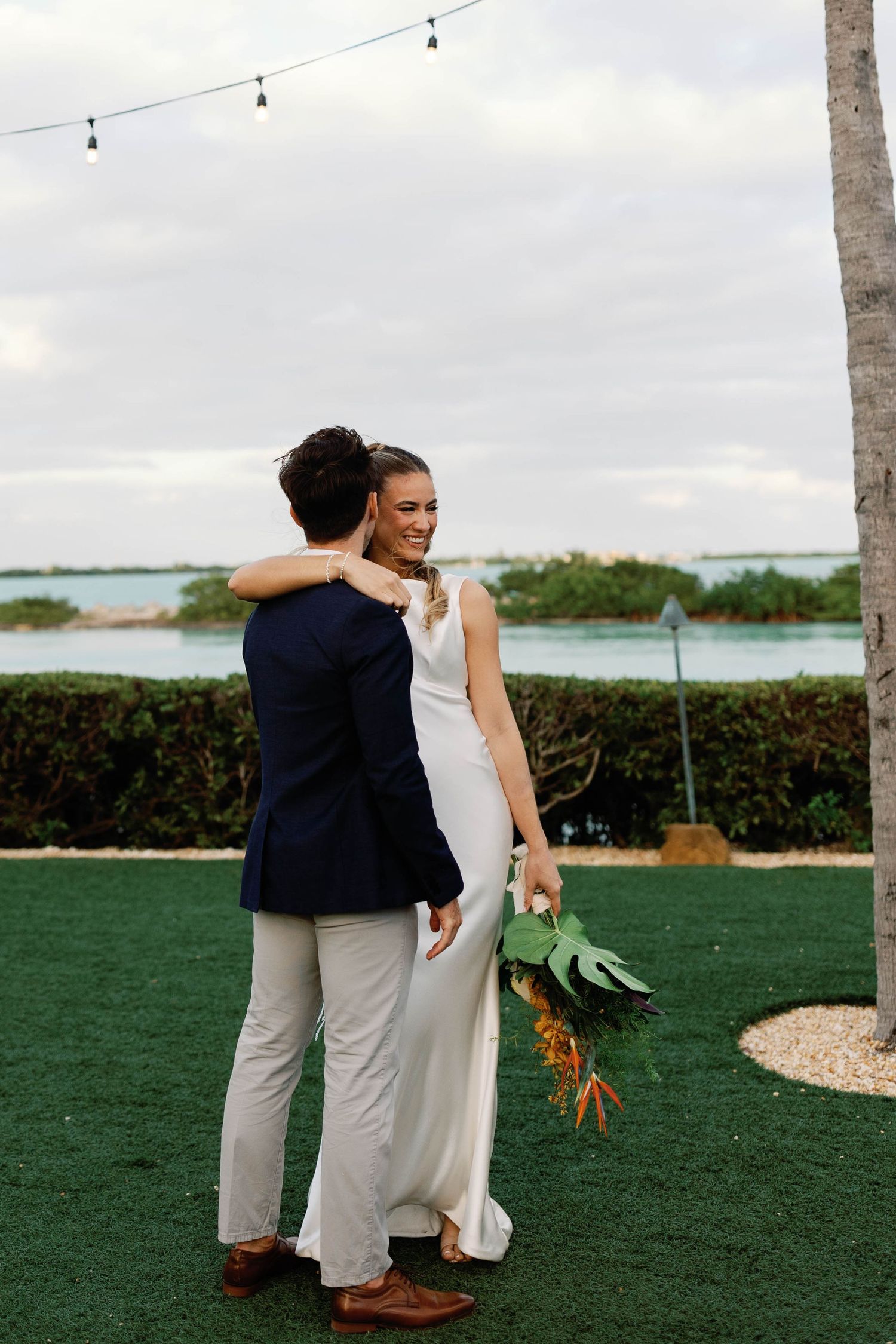 Wedding couple embraces on outdoor lawn with string lights and tropical landscaping.