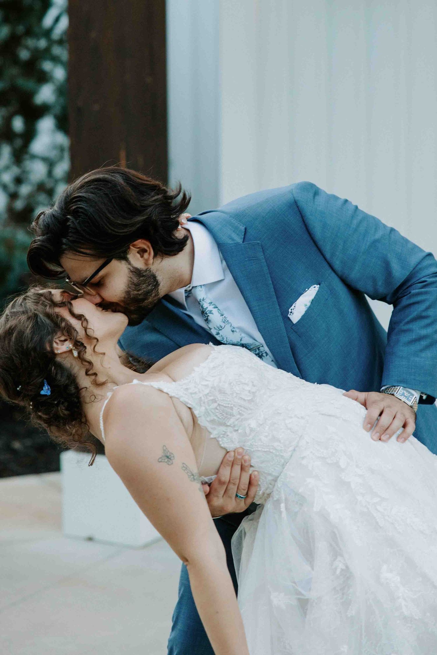 Romantic dip kiss between newlyweds with bride in lace dress and groom in blue suit.