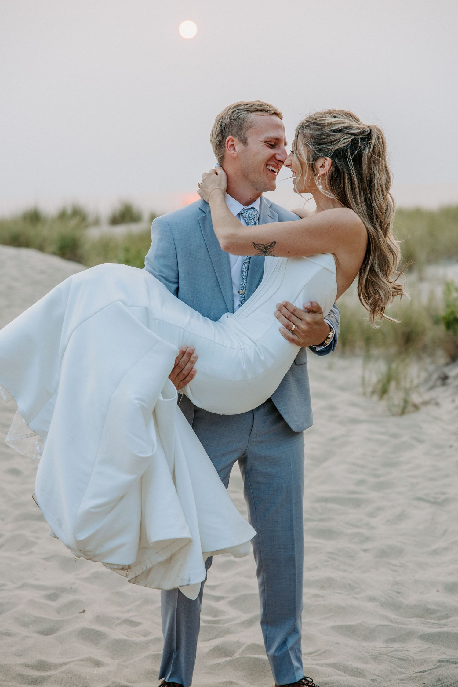 Romantic beach wedding photos with couple in blue suit and white dress against sandy dunes.