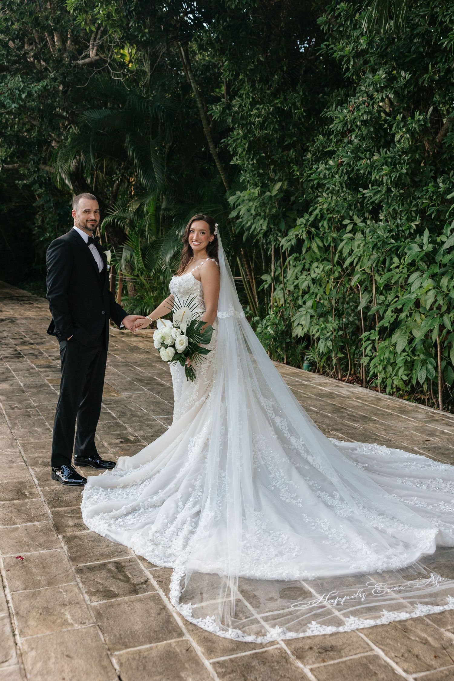 Wedding couple posing on stone patio showcasing bride's elaborate lace train and veil.