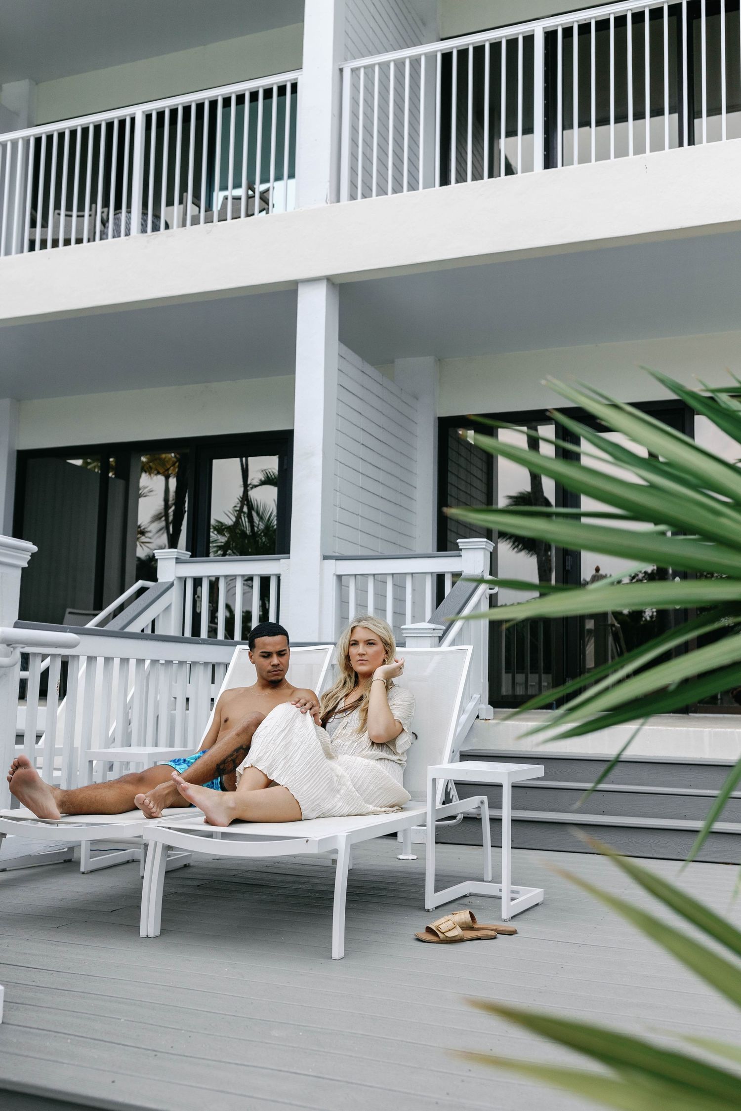 White lounge chairs on a modern resort patio with tropical plants and railings in the background.